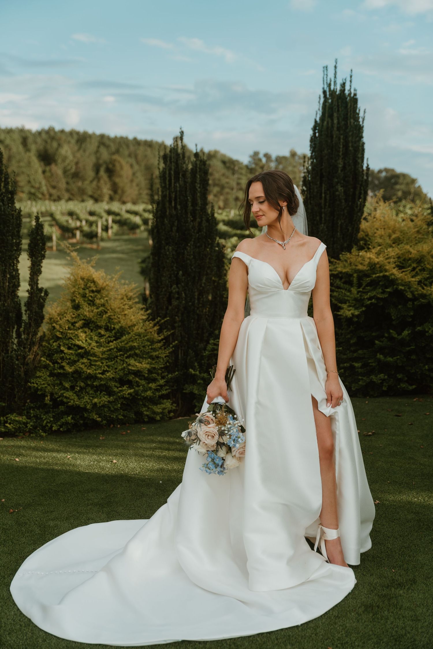 Bride in elegant off-shoulder white ball gown with train holding bouquet in lush garden with cypress trees.