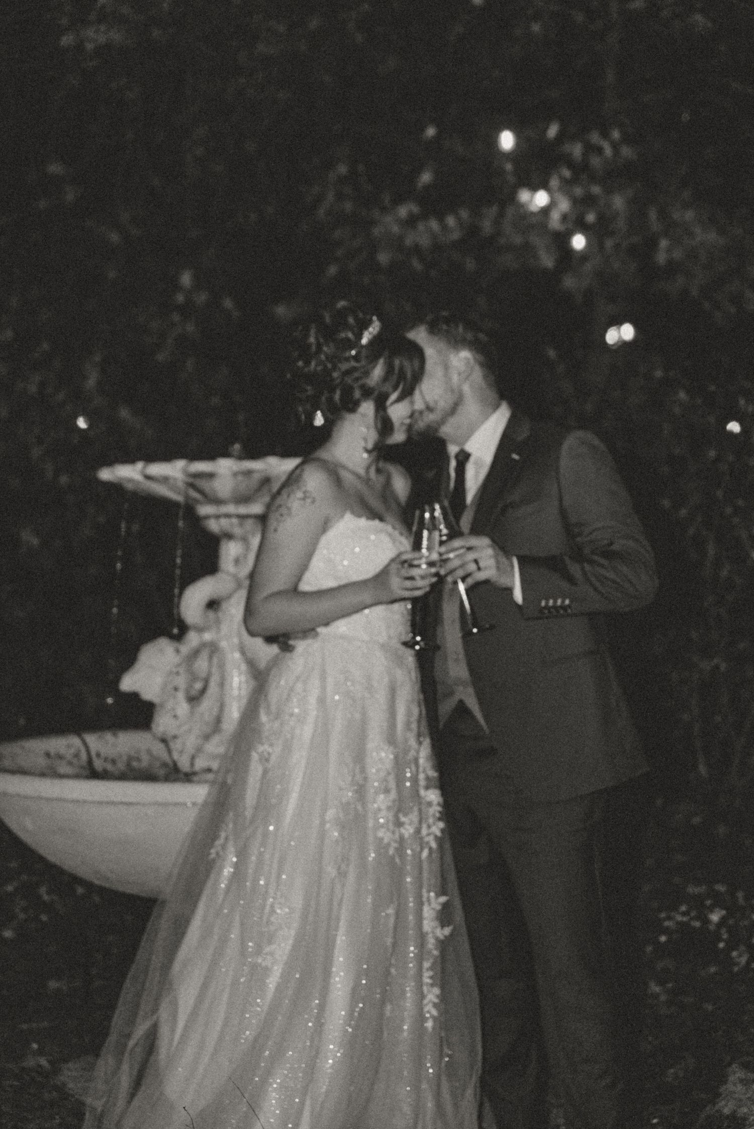 Black and white romantic wedding photo of bride and groom sharing a moment by a garden fountain at night.