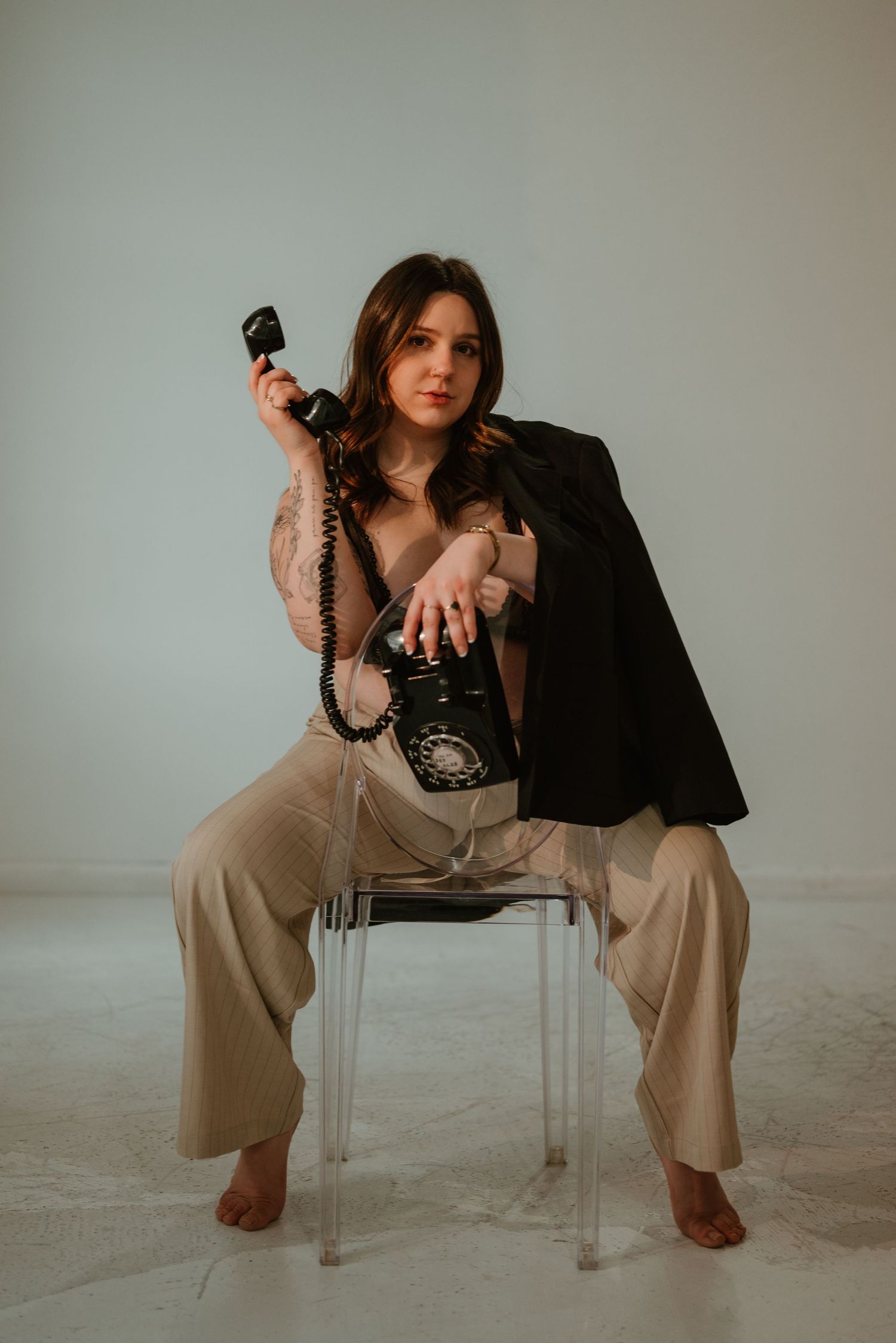 Barefoot woman in beige wide-leg pants holds retro rotary phone handset on clear acrylic chair.
