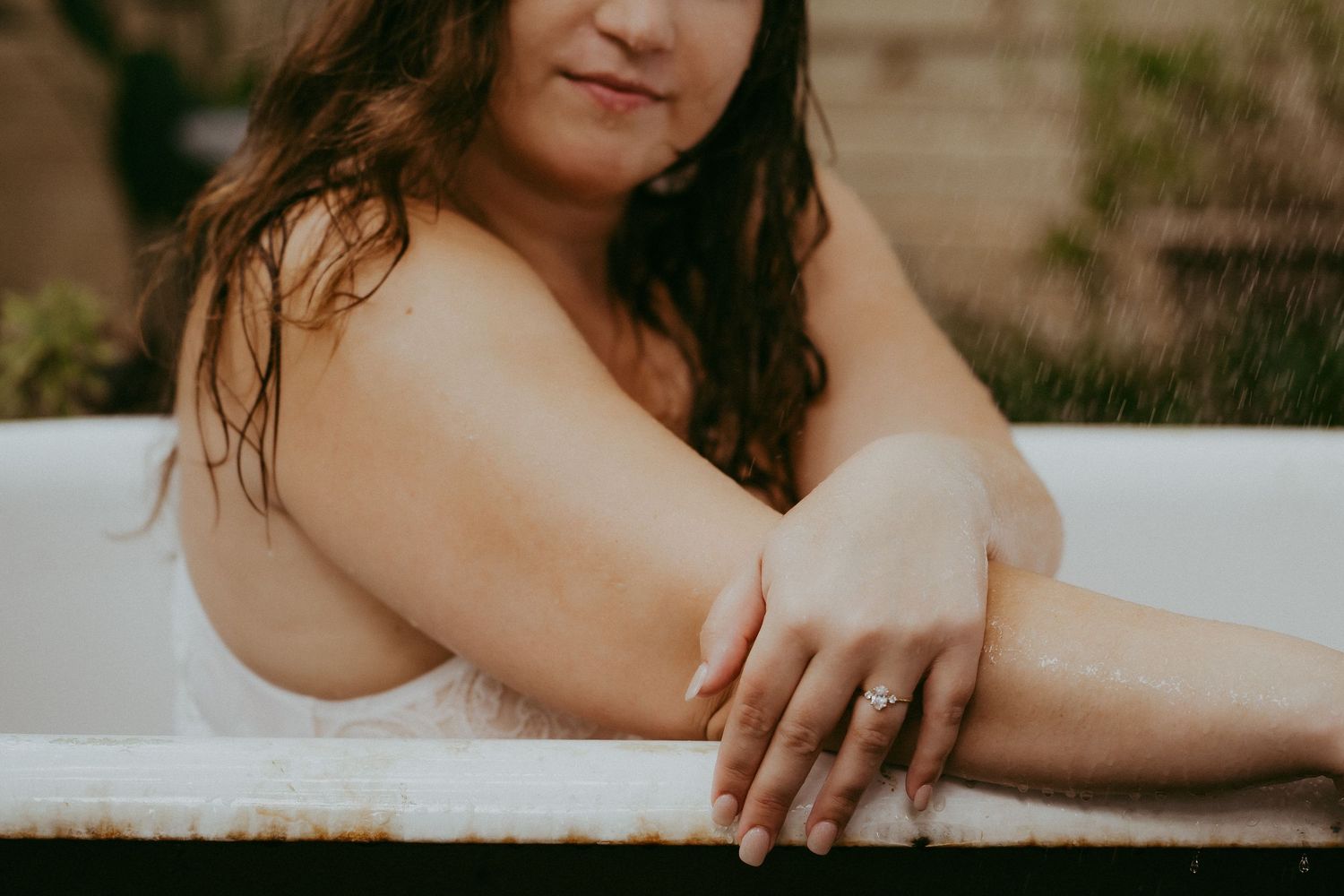A brunette woman relaxes in an outdoor bathtub, arms resting on the edge, wearing a ring, looking at camera.