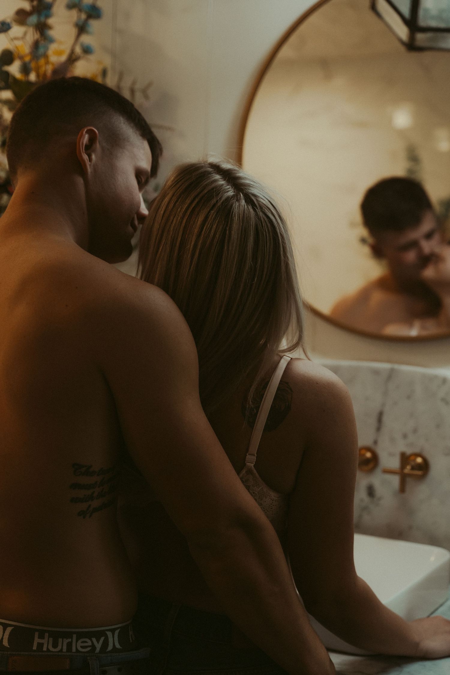 Couple sharing intimate moment together near marble bathroom sink with round mirror reflection.