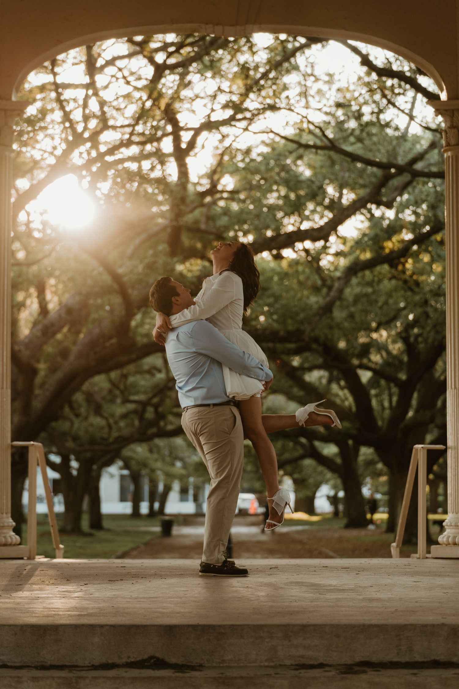 Couple sharing a romantic lift embrace under a sunlit archway surrounded by oak trees.