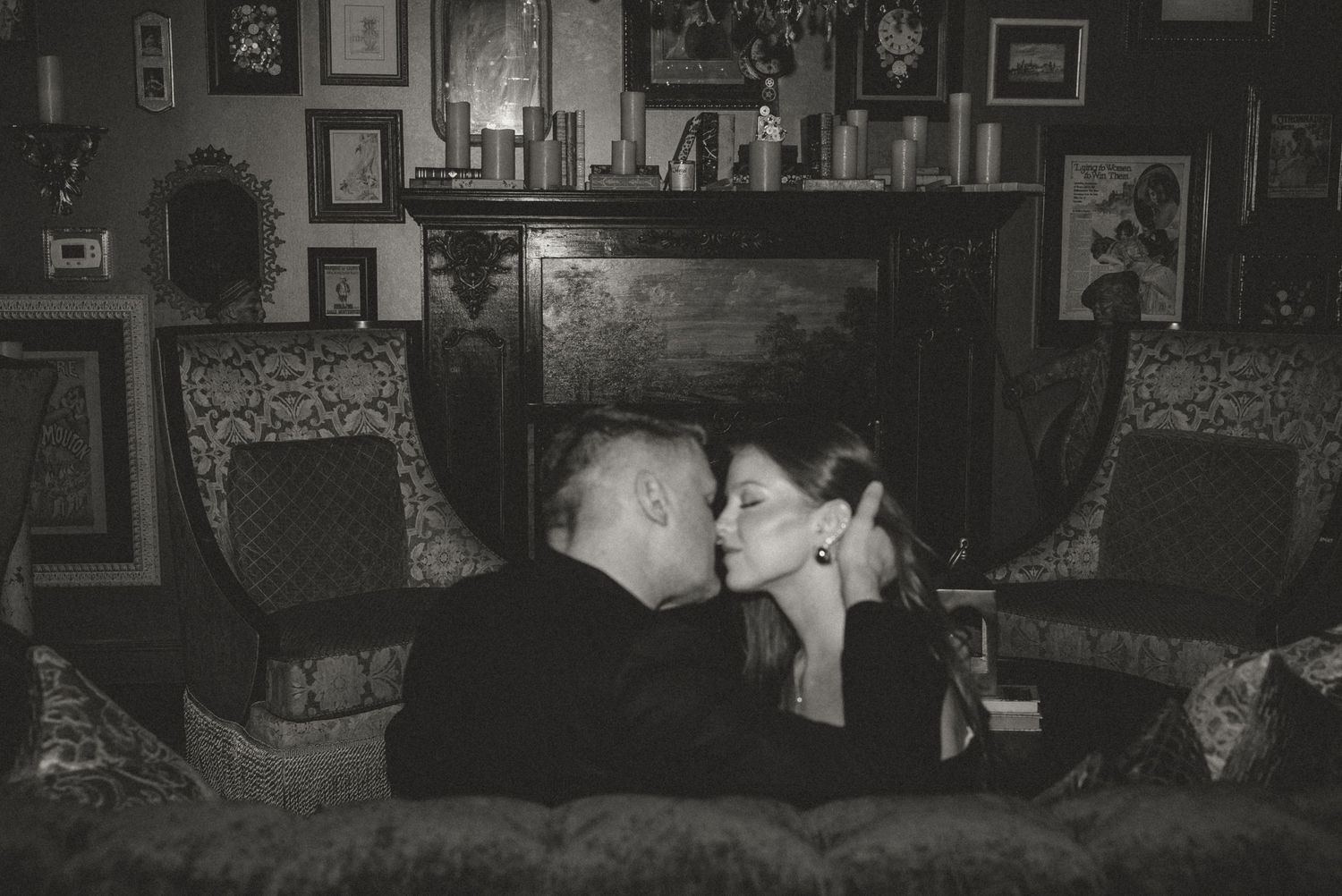 Black and white photo of a couple sharing a kiss on a couch by a fireplace in an elegant room.