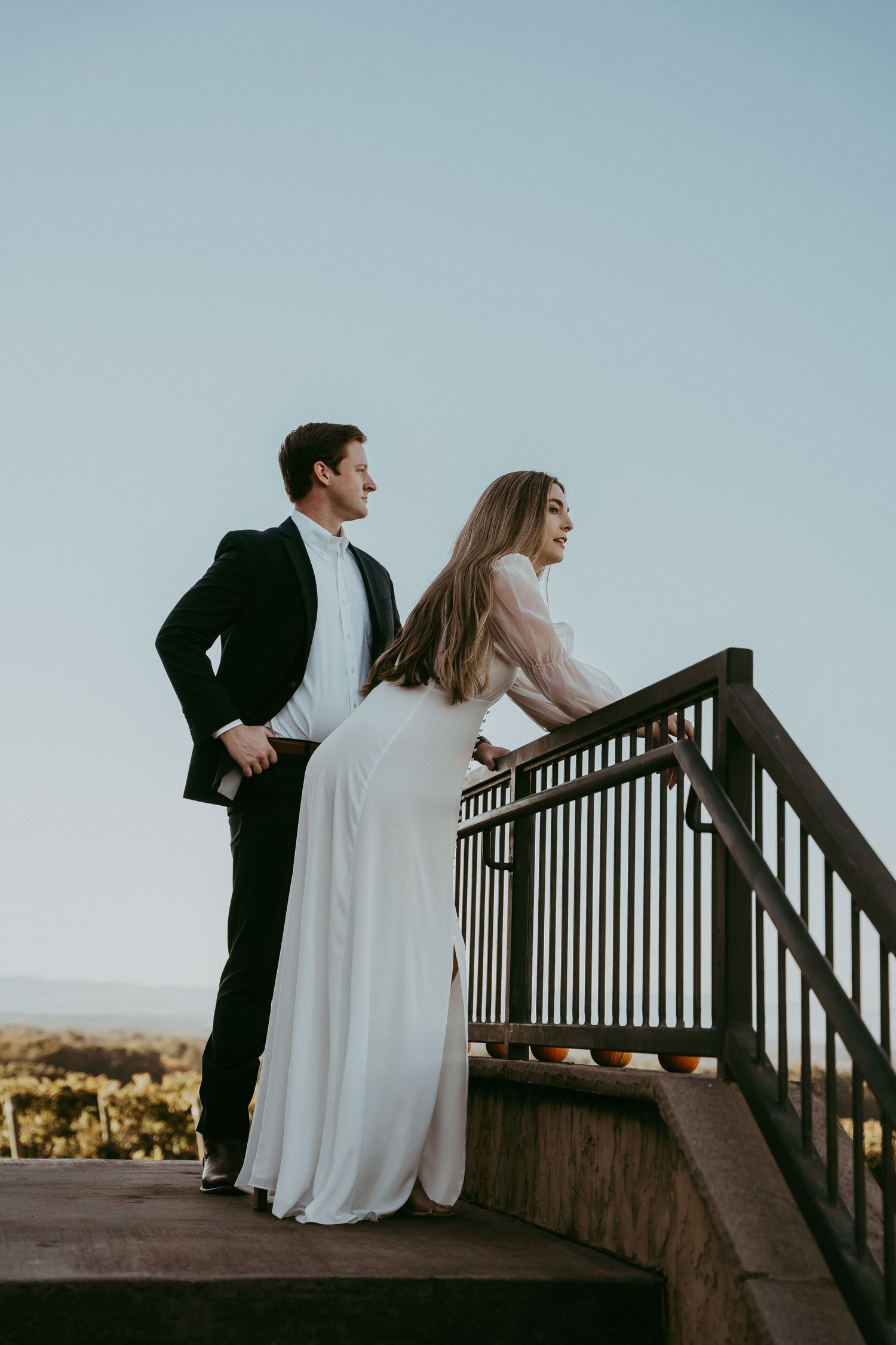 Elegant couple in wedding attire standing on a staircase overlooking a scenic landscape.