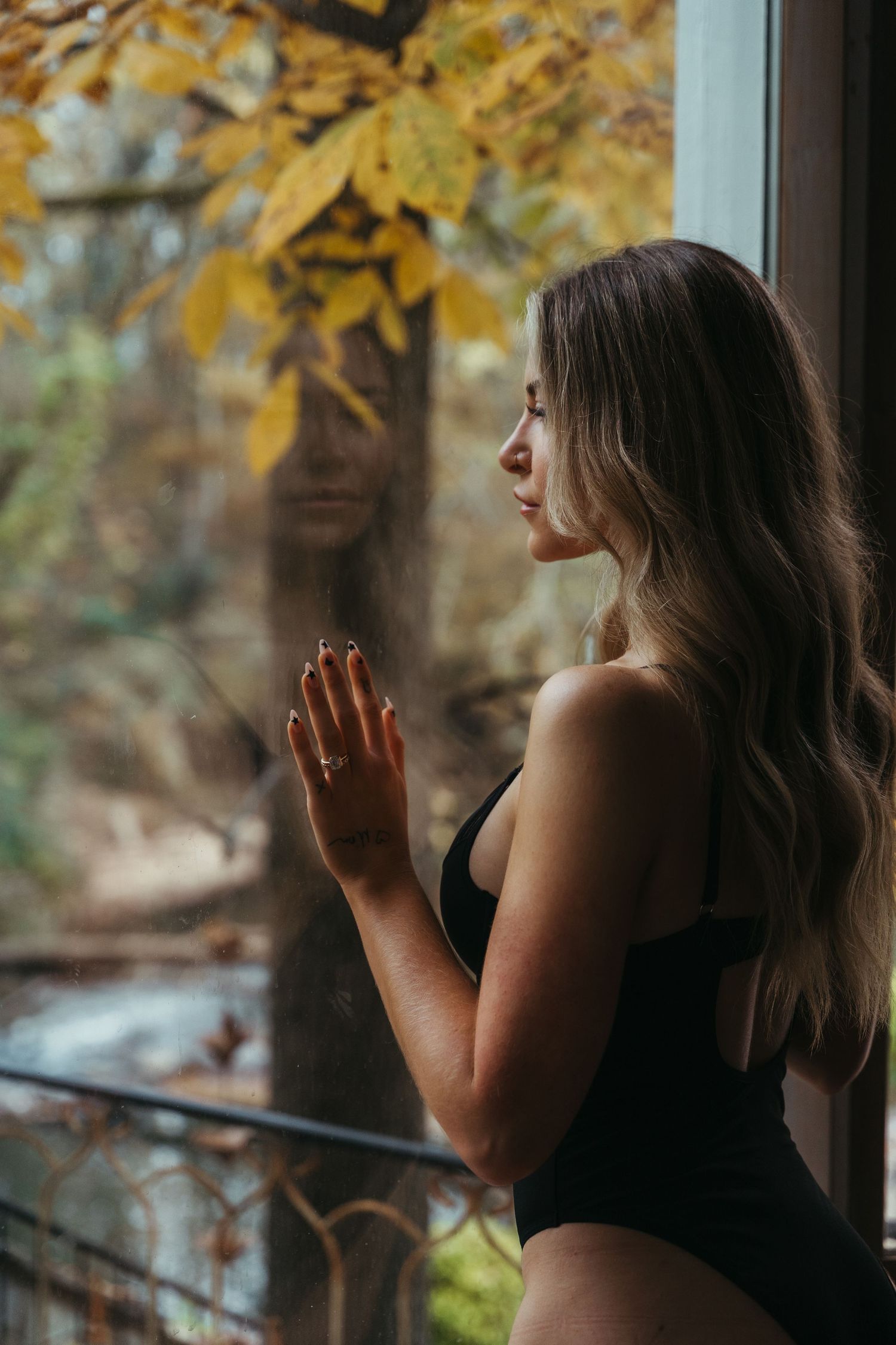 Blonde woman in black bodysuit touching glass window with fall foliage outside.