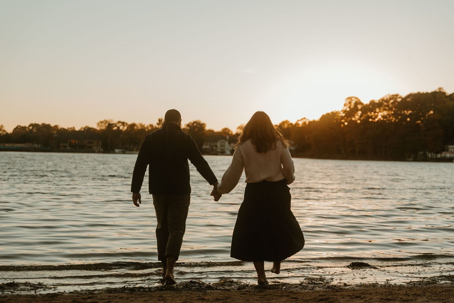 Couple holding hands walking along a serene lakeshore at beautiful golden sunset.
