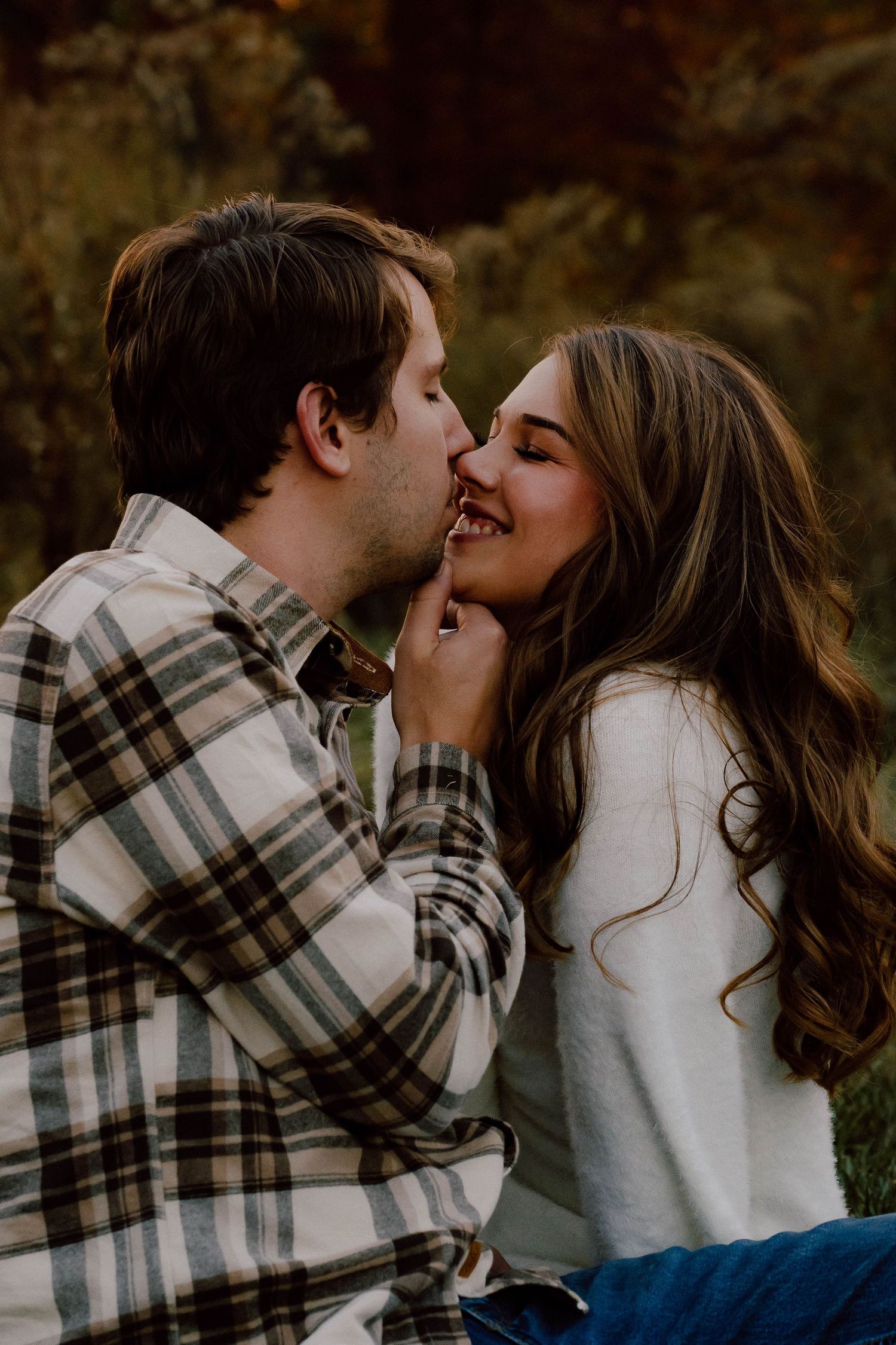 Couple sharing a playful kiss outdoors in fall, man in plaid shirt, woman in white sweater.