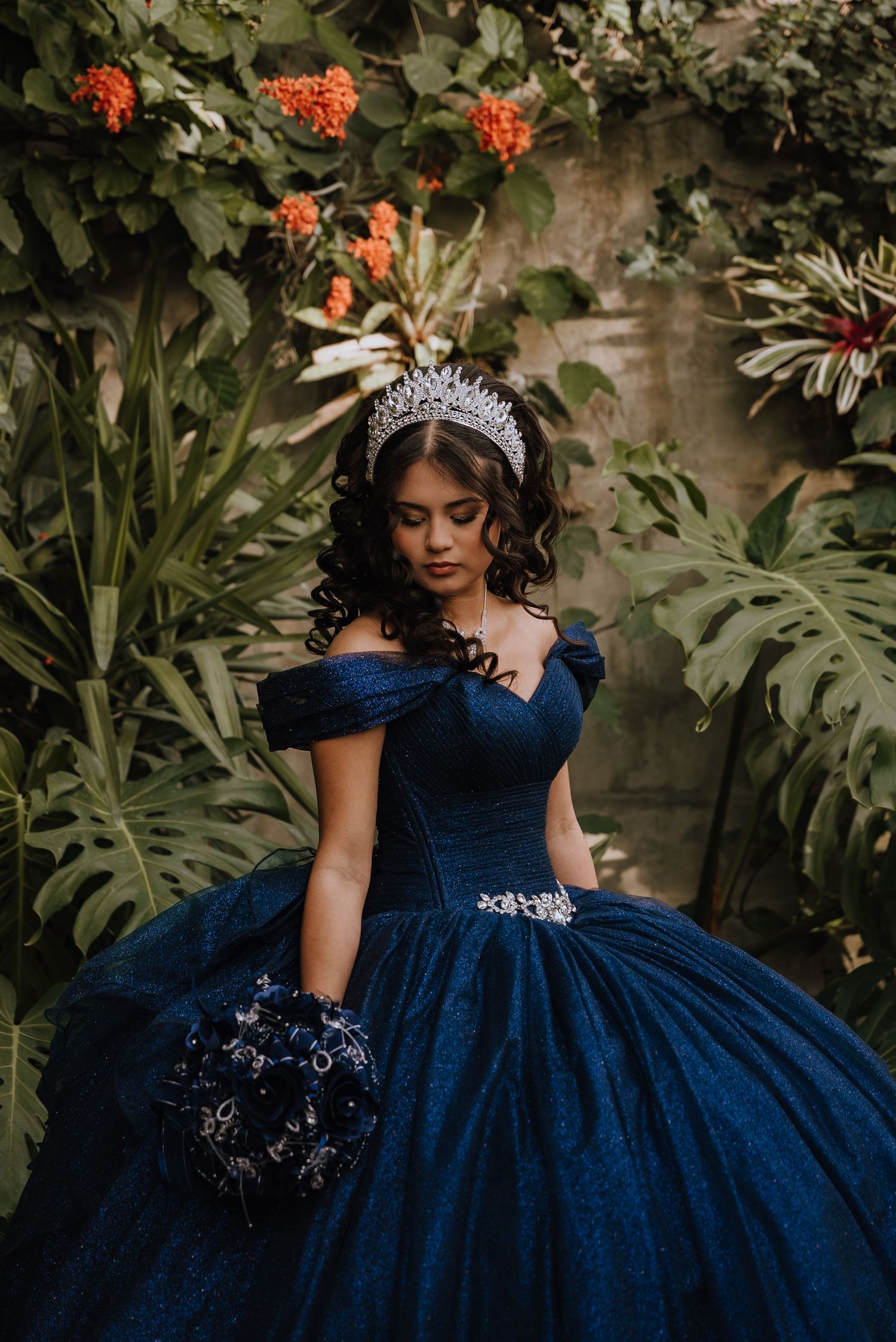 A young woman in a navy blue quinceañera gown and crystal crown holds a bouquet among tropical plants.