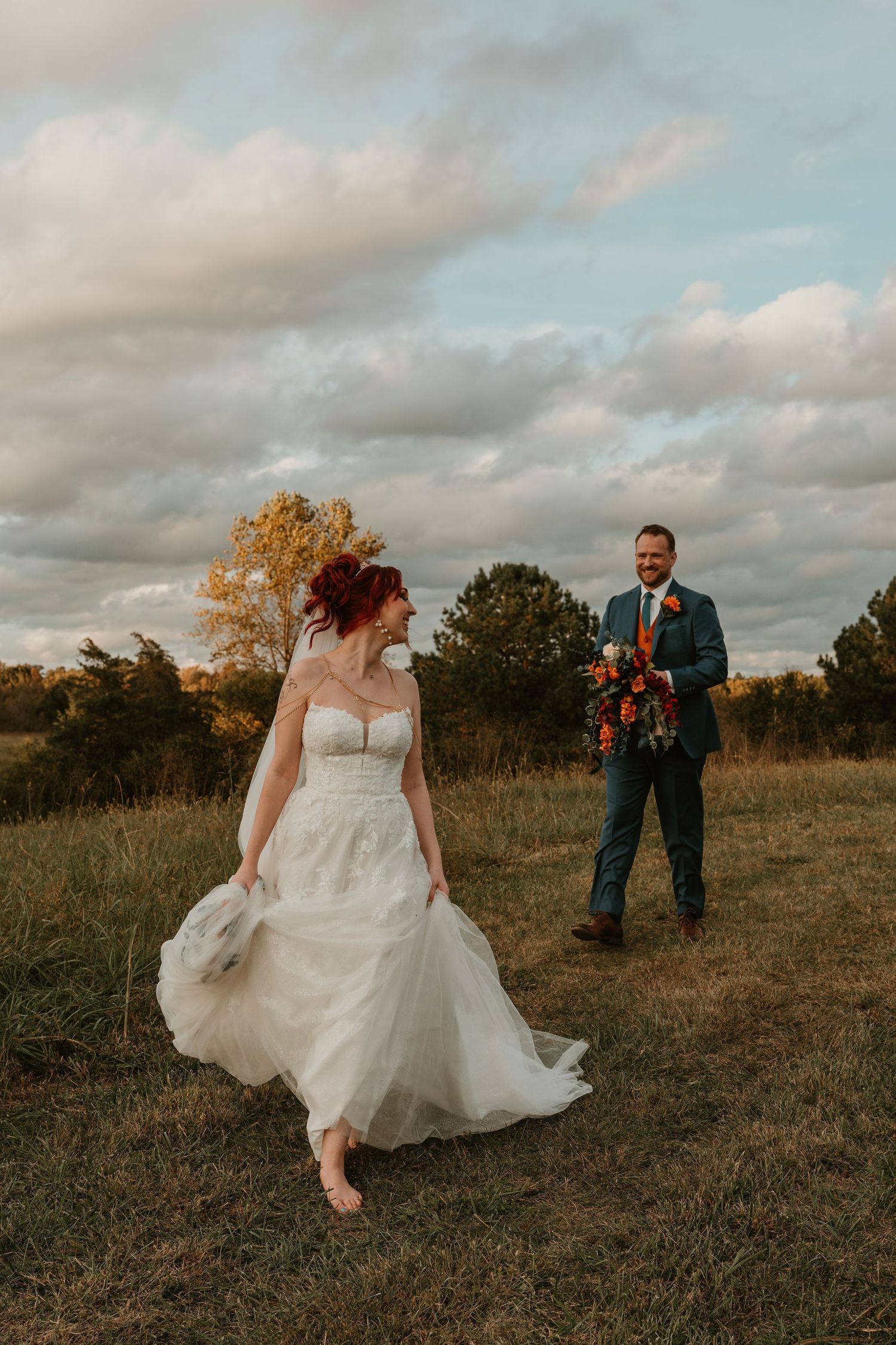 Bride in white gown walks through autumn field while groom in blue suit holds bouquet at golden hour.