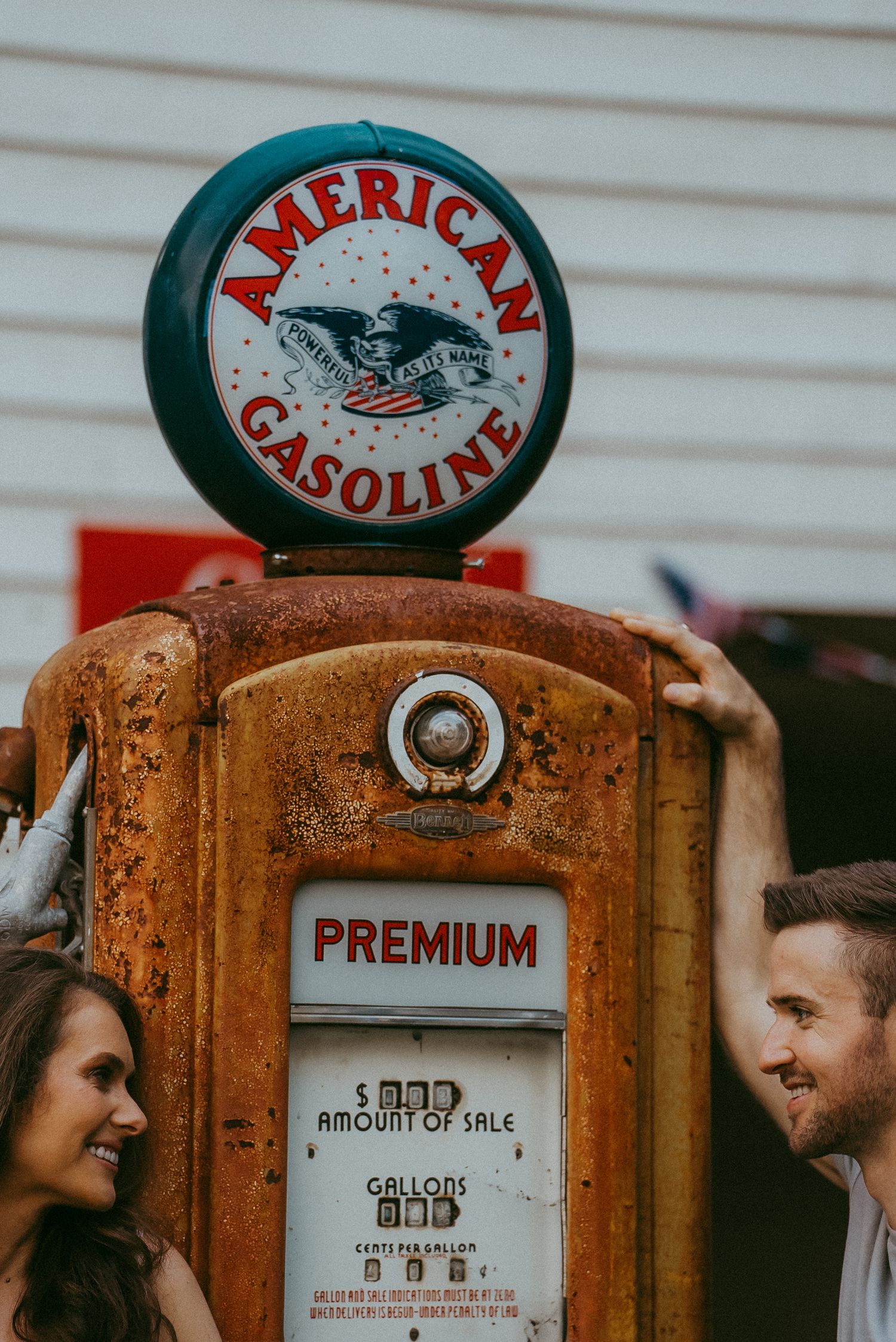 Couple smiling beside a rusty vintage American Gasoline premium pump during an outdoor photo shoot.