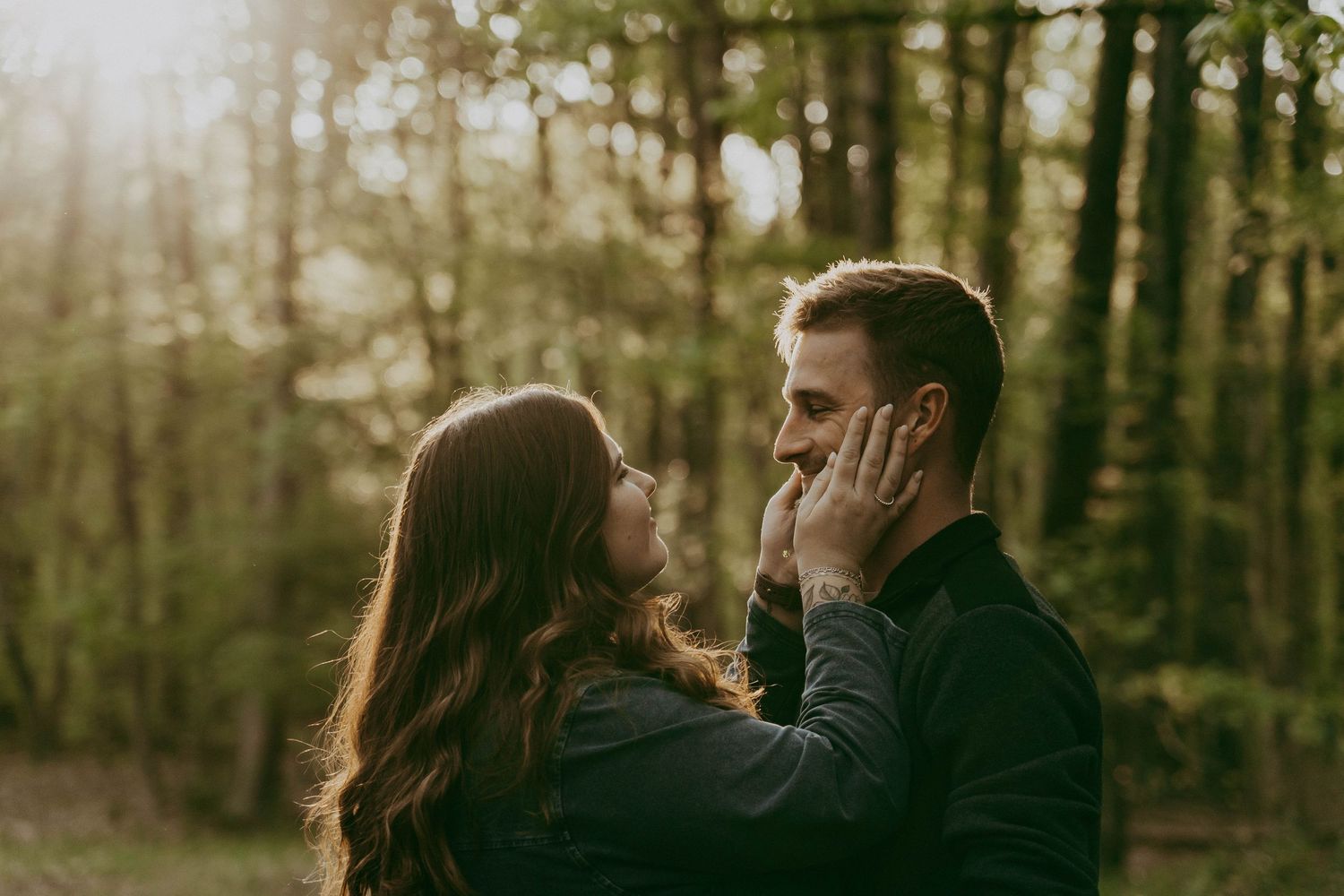 Couple sharing a tender moment in a sunlit forest, woman gently holding man's face.