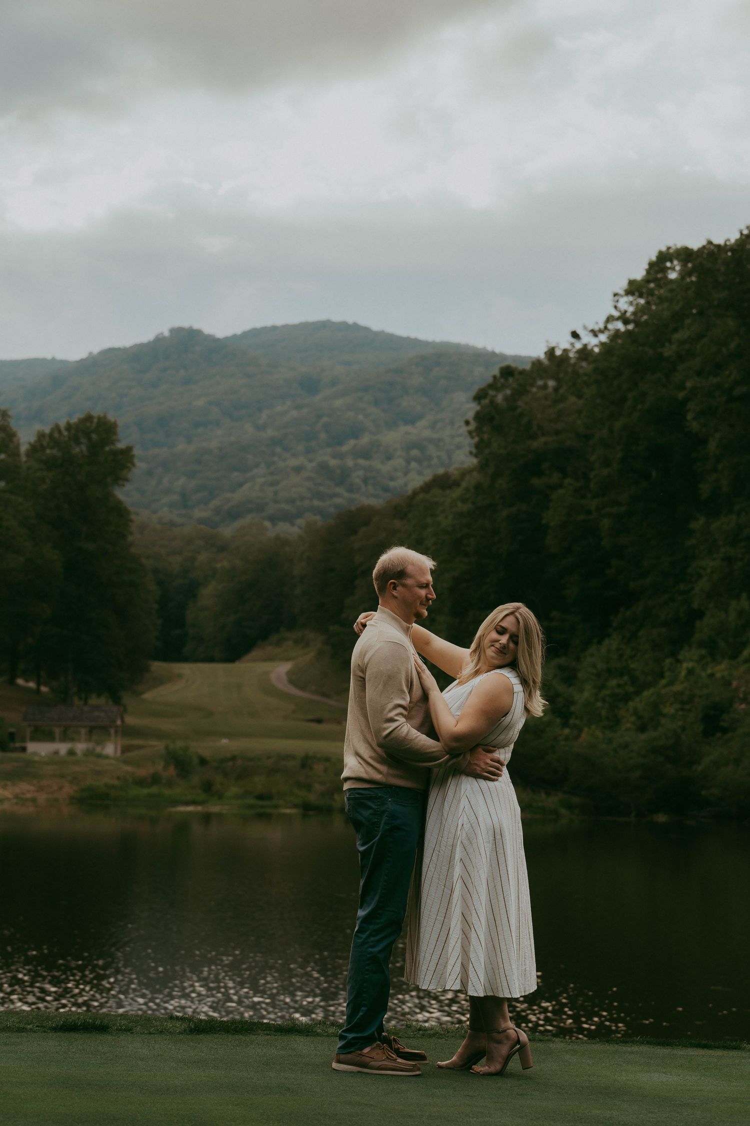 Couple embracing on a green lawn with a serene mountain lake and cloudy sky backdrop.