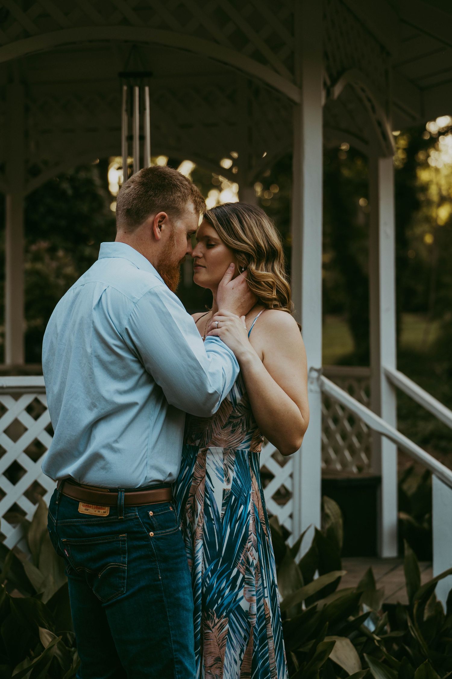 Couple sharing an intimate almost-kiss near a white gazebo in a lush garden at sunset.