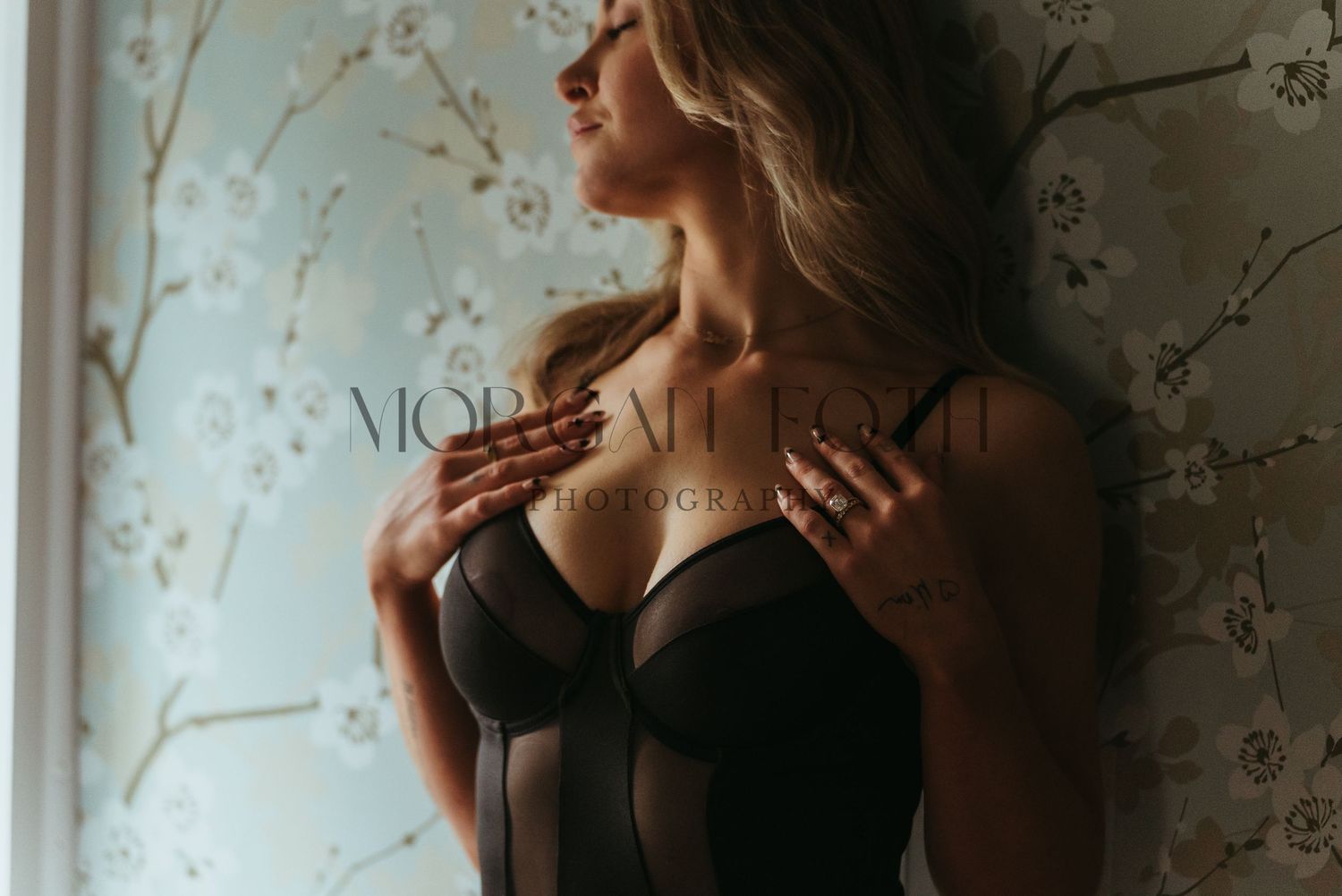 Close-up of woman in black lingerie posing near window with floral wallpaper.