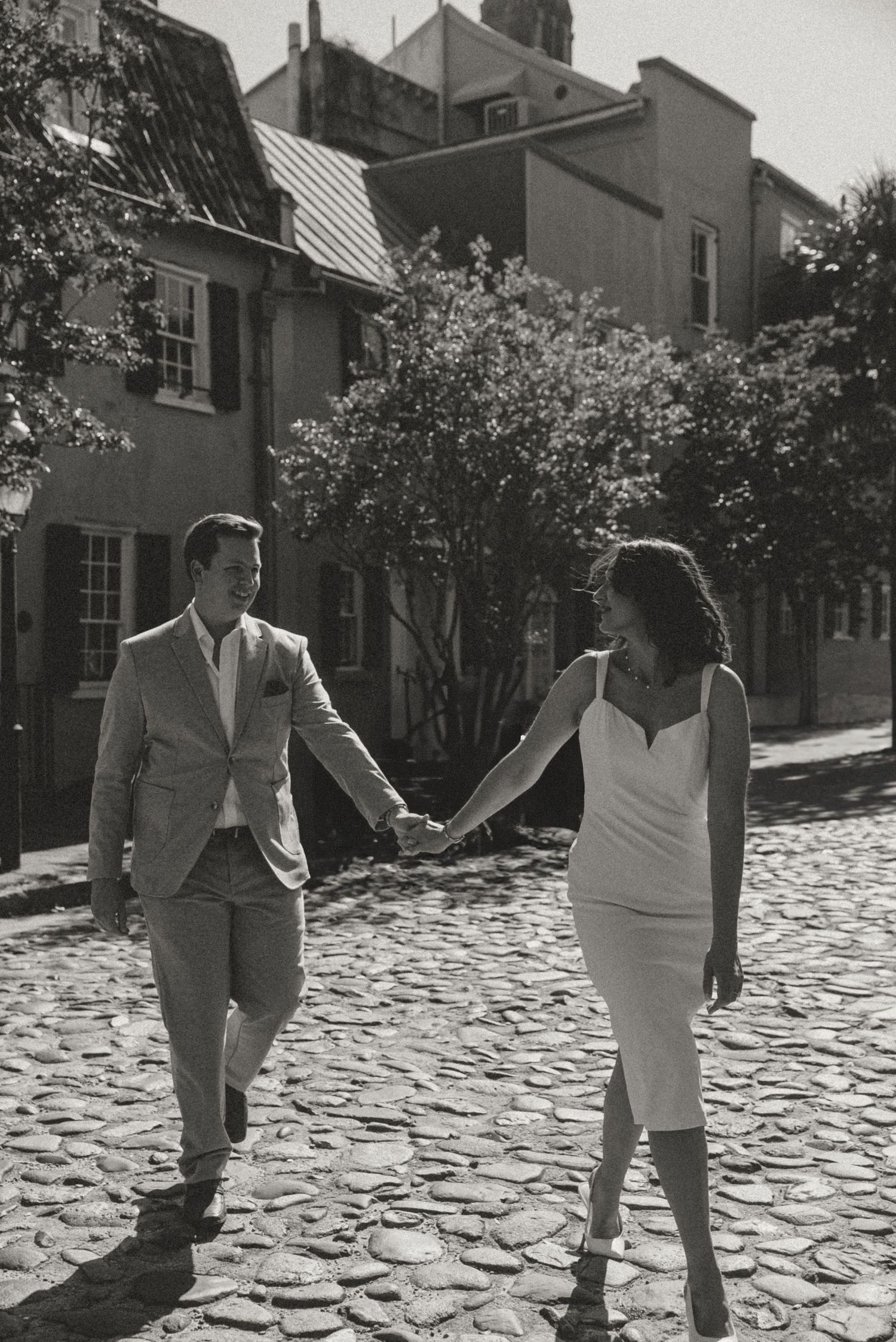 Black and white photo of couple holding hands walking on cobblestone street.