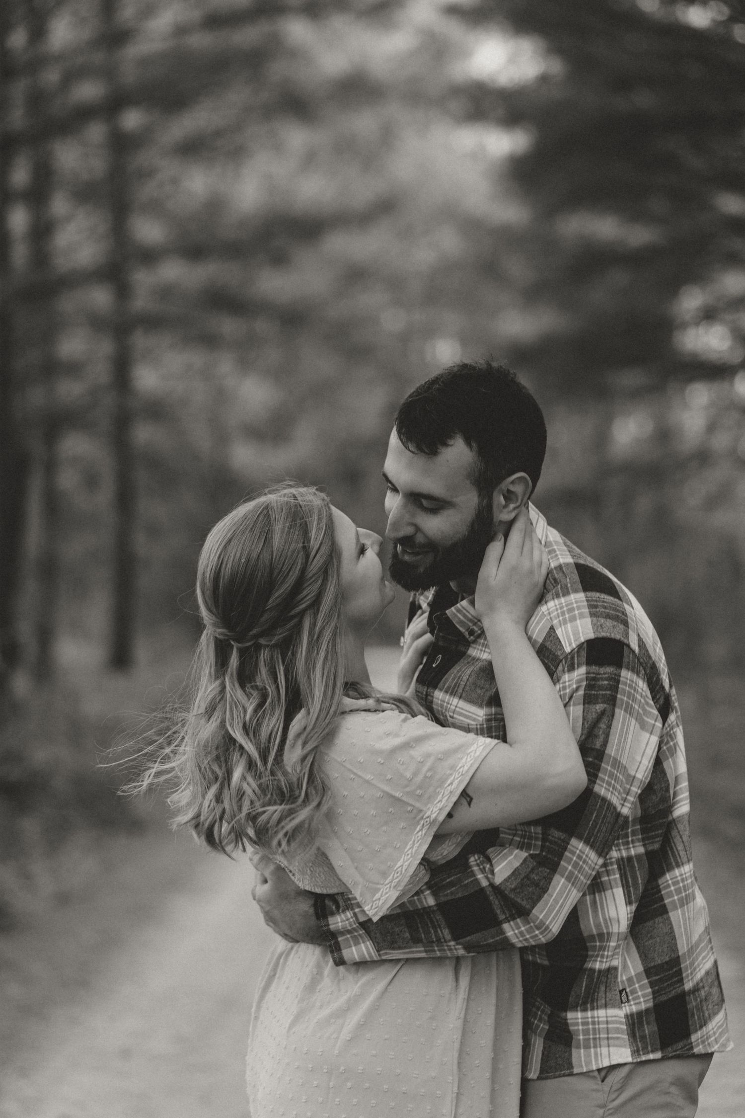 Black and white photo of a couple embracing and almost kissing on a forest path.