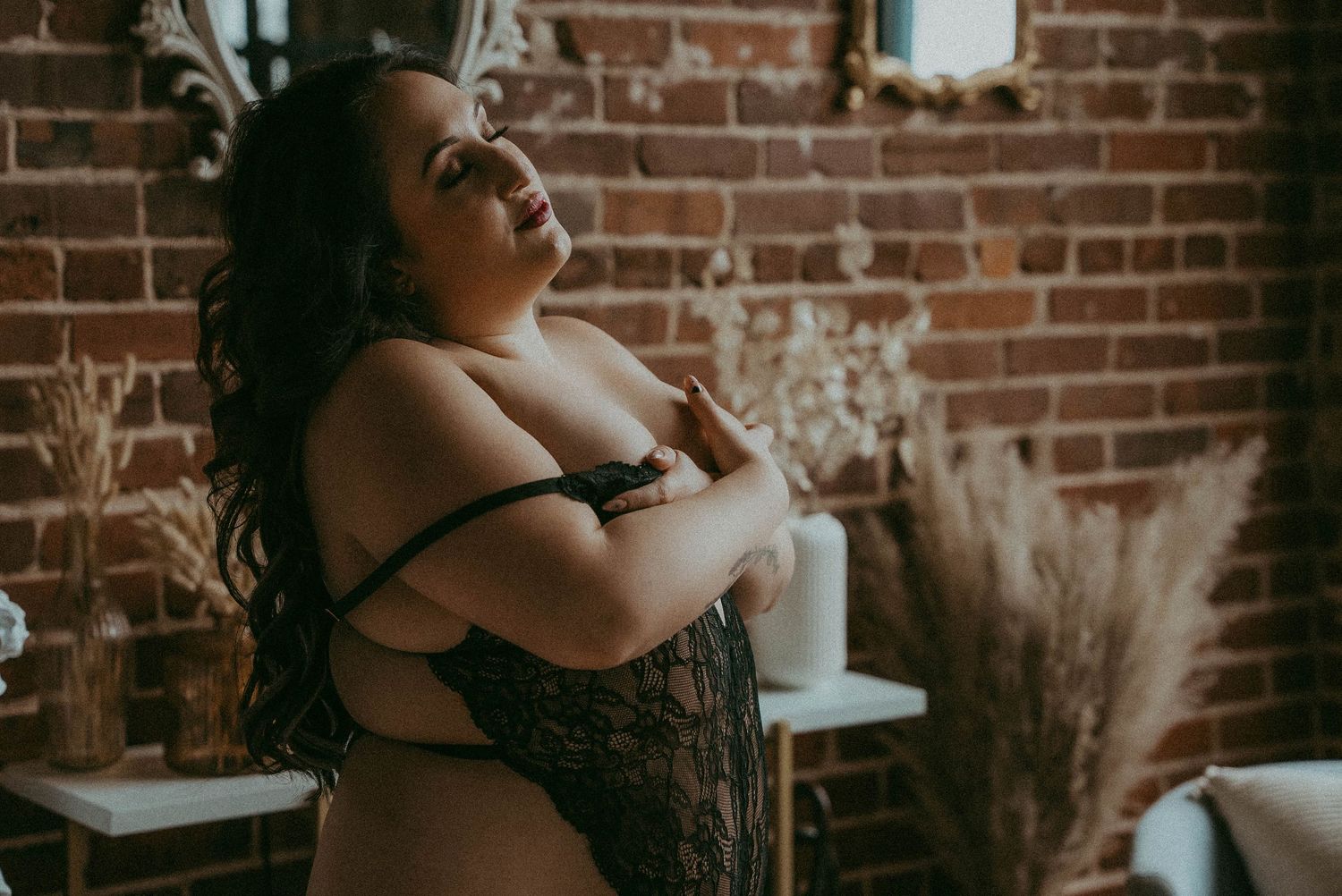 A plus-size woman in black lace lingerie poses confidently in front of a rustic brick wall with dried florals.