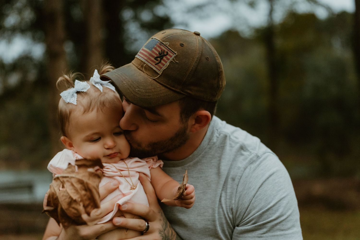 A father in a camo cap lovingly kisses his baby girl with a bow in her hair outdoors.