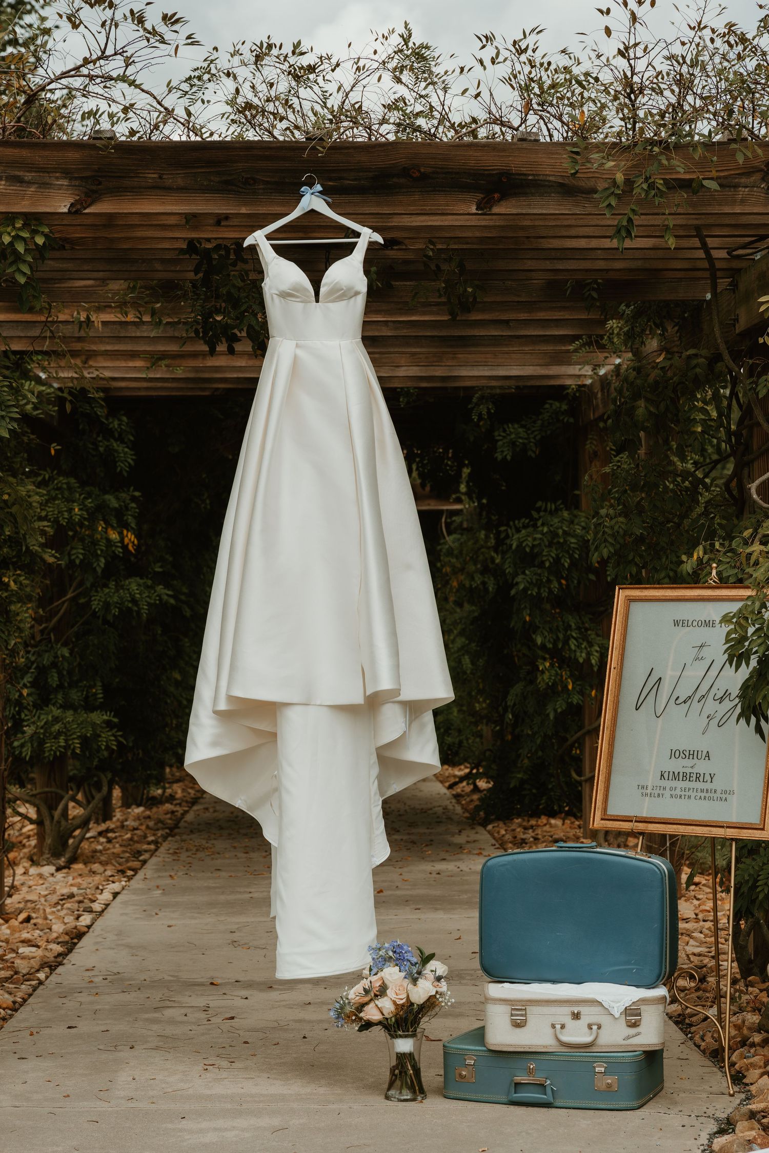 White ball gown wedding dress hanging outdoors beside vintage blue suitcases and welcome sign with bouquet.