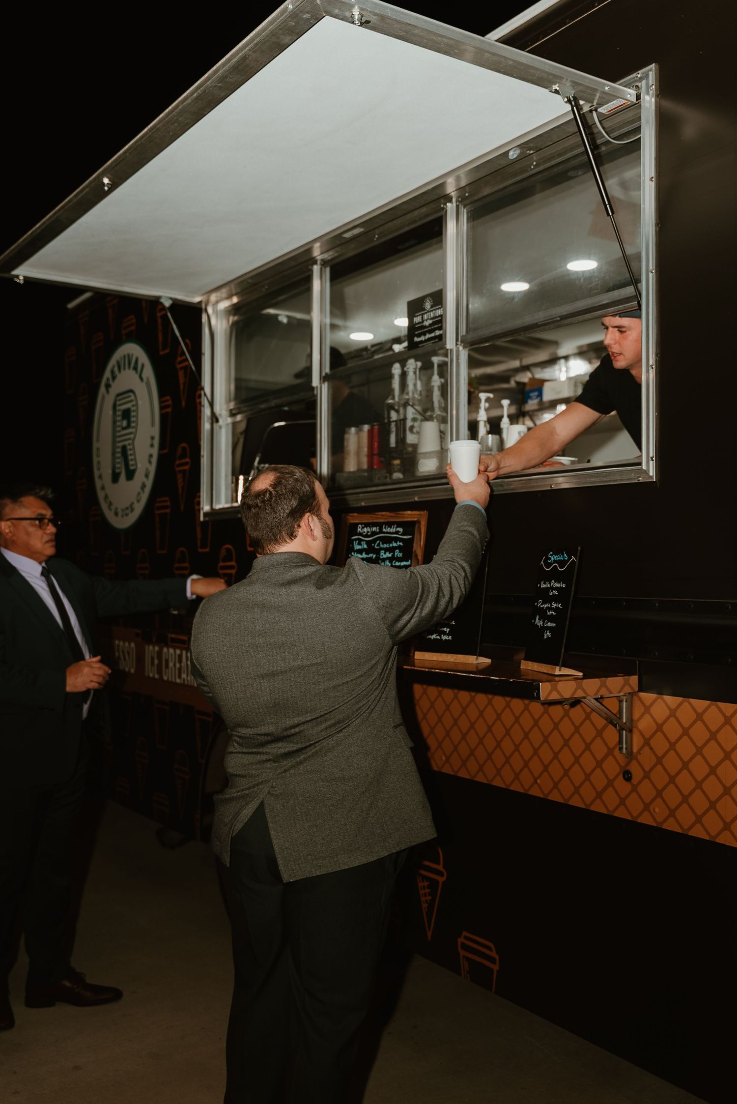 Wedding guests ordering from a dark food truck at night with server handing items through window.