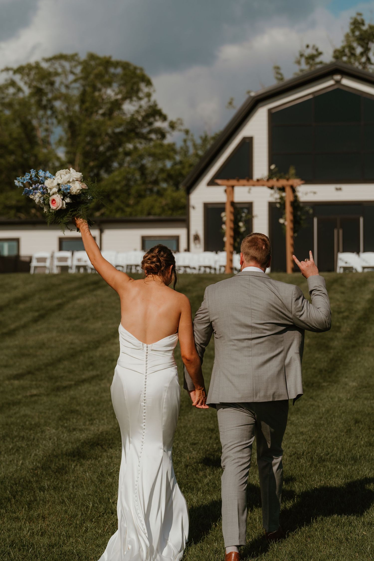 Bride and groom walk hand-in-hand toward a white barn wedding venue, raising their hands in celebration.