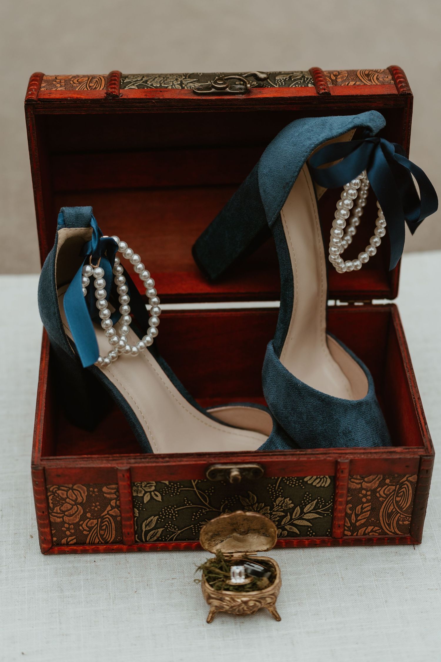 Blue velvet heels with pearl ankle straps displayed in ornate wooden treasure chest with wedding rings.