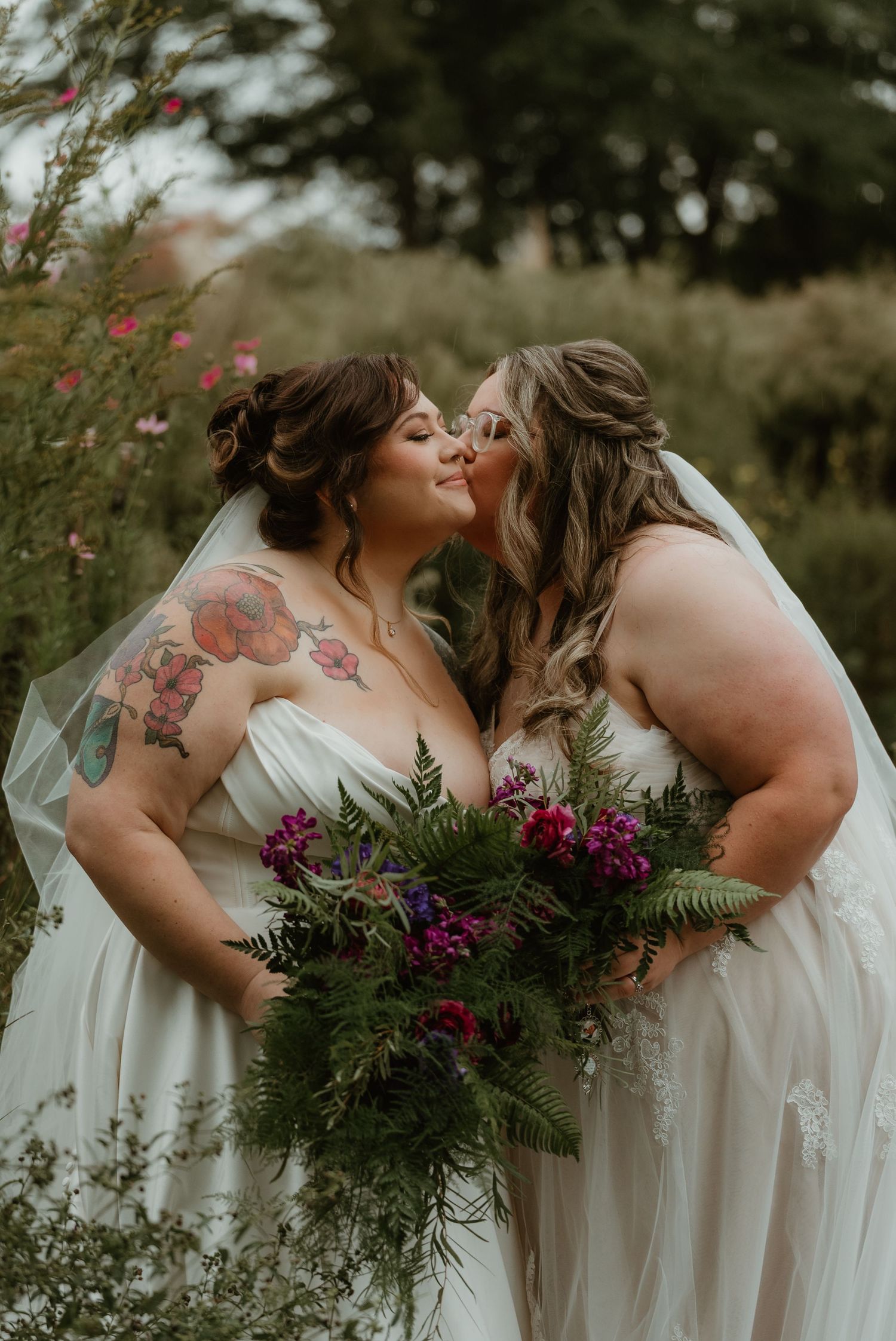 Two brides in white gowns share a kiss surrounded by lush greenery and vibrant floral bouquets.