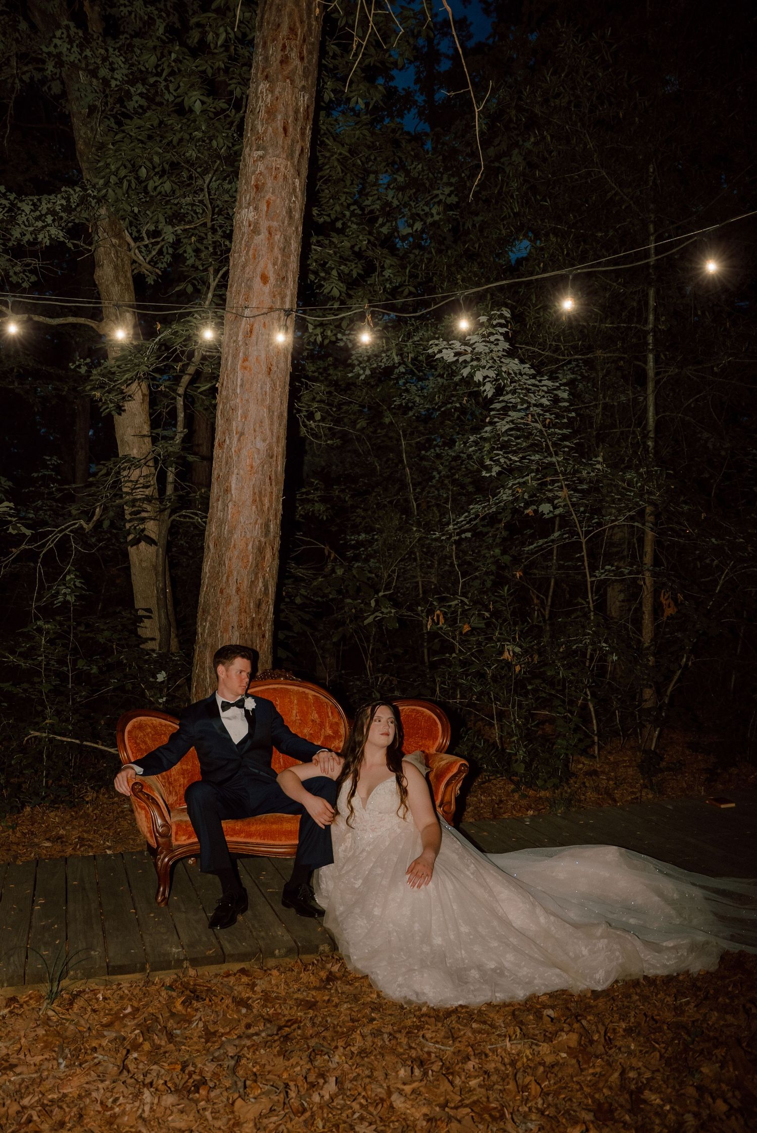Bride and groom seated on vintage orange velvet sofa under string lights in romantic forest at night.