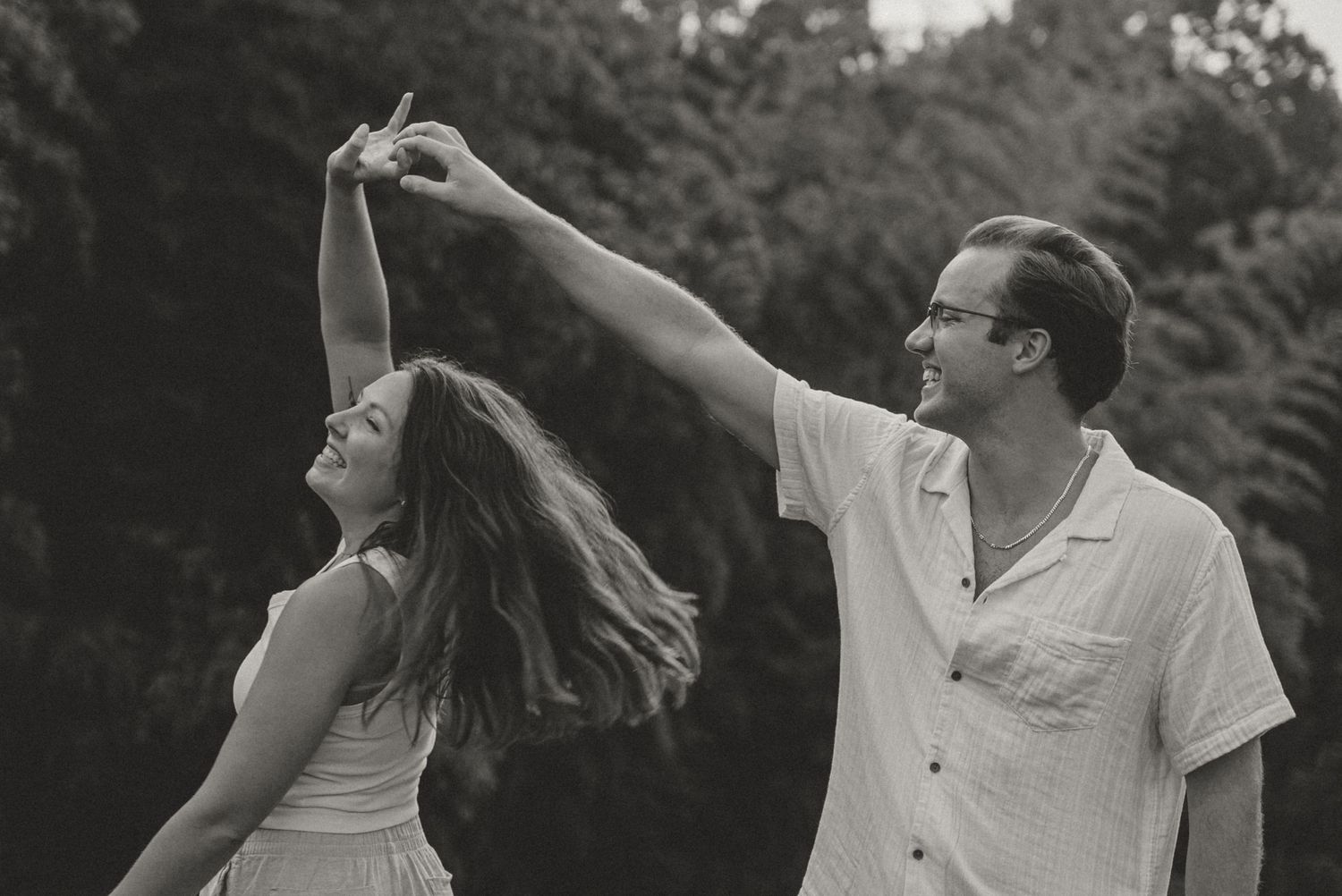 Black and white photo of a man spinning a woman outdoors with trees in the background during an engagement shoot.