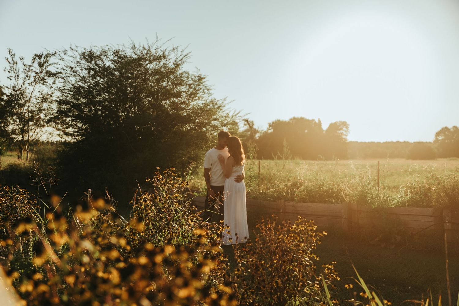 Couple kissing among wildflowers in a sunlit field surrounded by lush golden hour scenery.