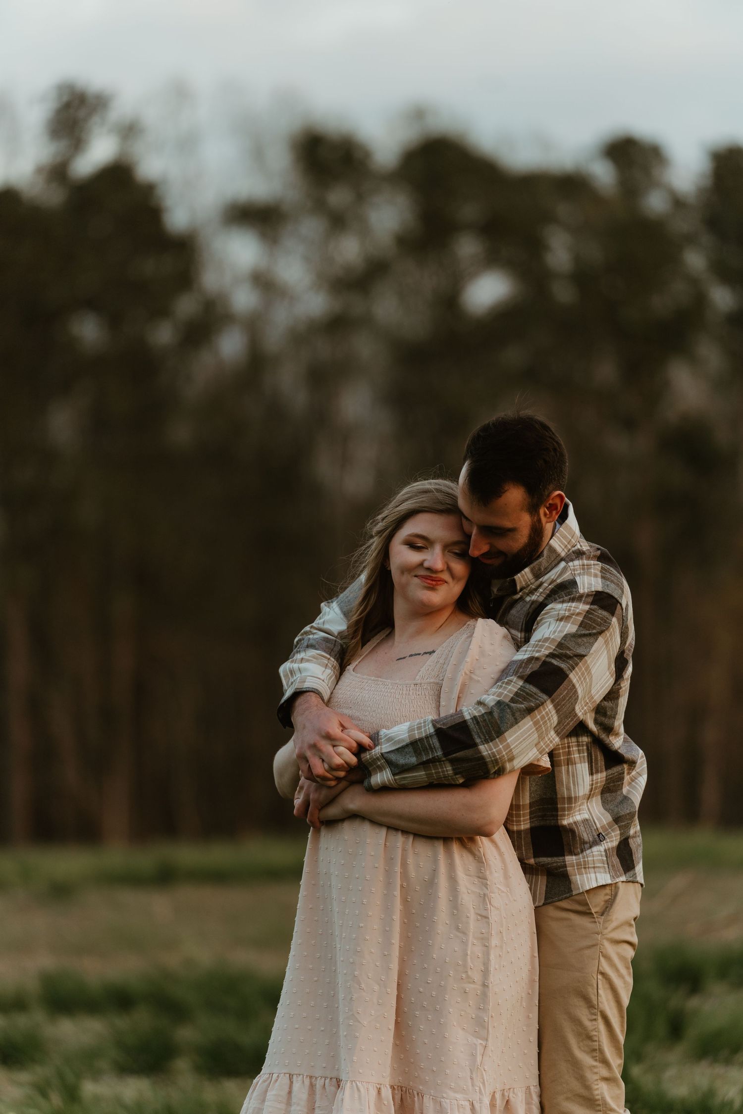 Man in plaid shirt embracing woman in pink dress in a golden field at sunset.