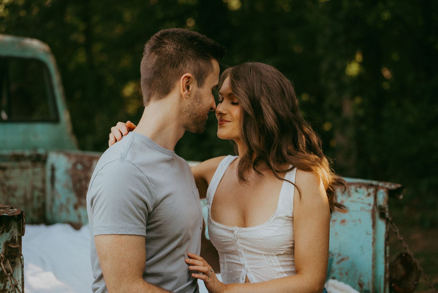 Couple sharing an intimate moment near a rustic turquoise vintage truck surrounded by green trees.