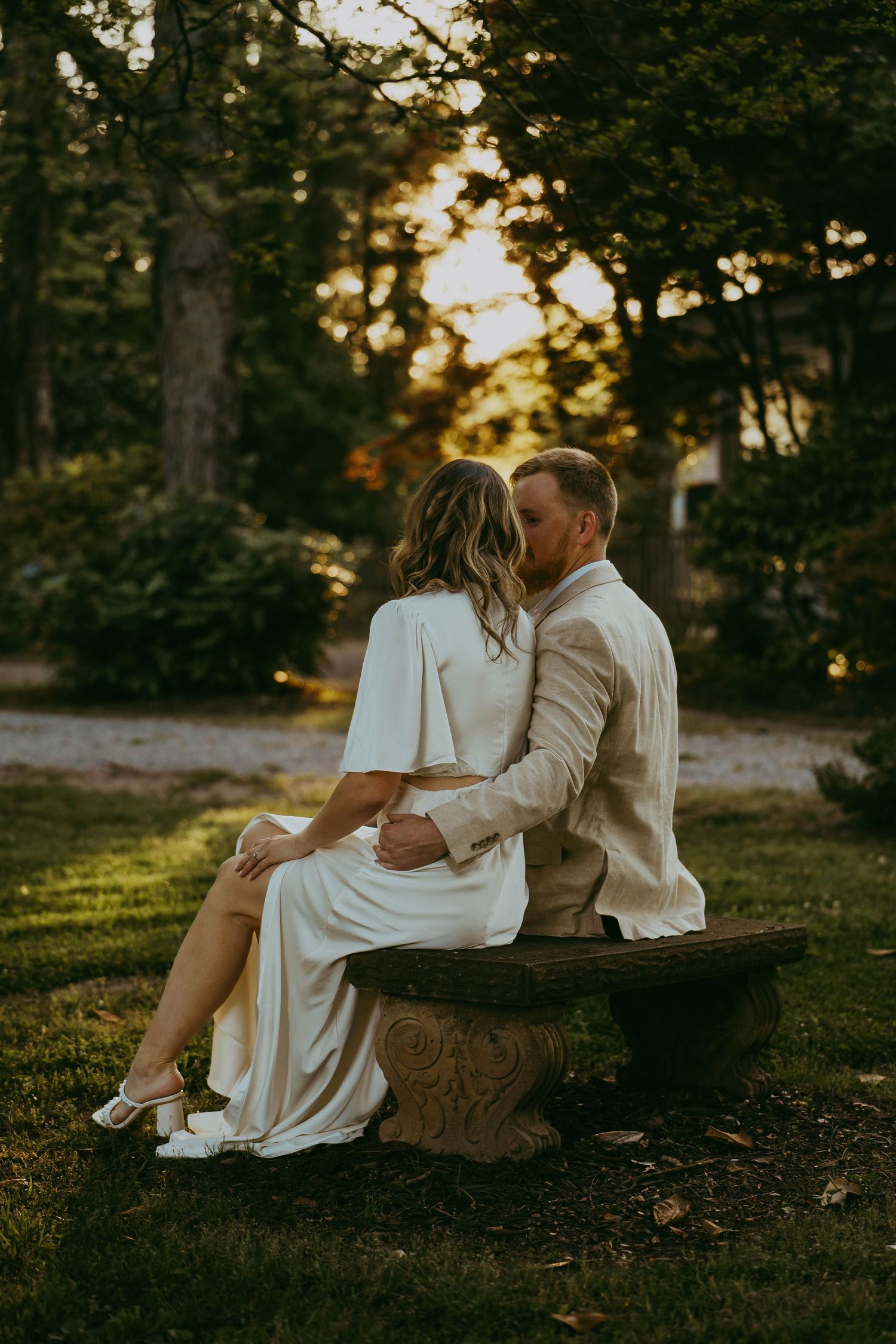Couple sitting closely on a stone bench sharing a kiss in a golden sunset garden.