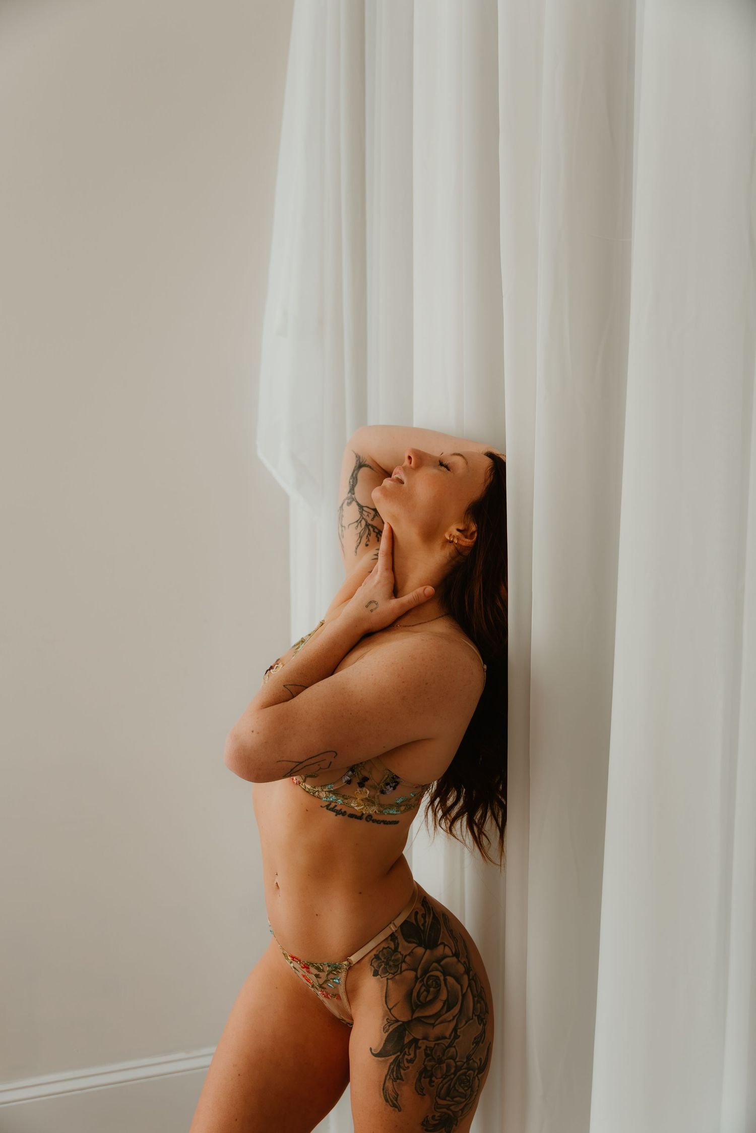 Tattooed woman in green bikini leaning against white curtains with head tilted back in boudoir portrait.