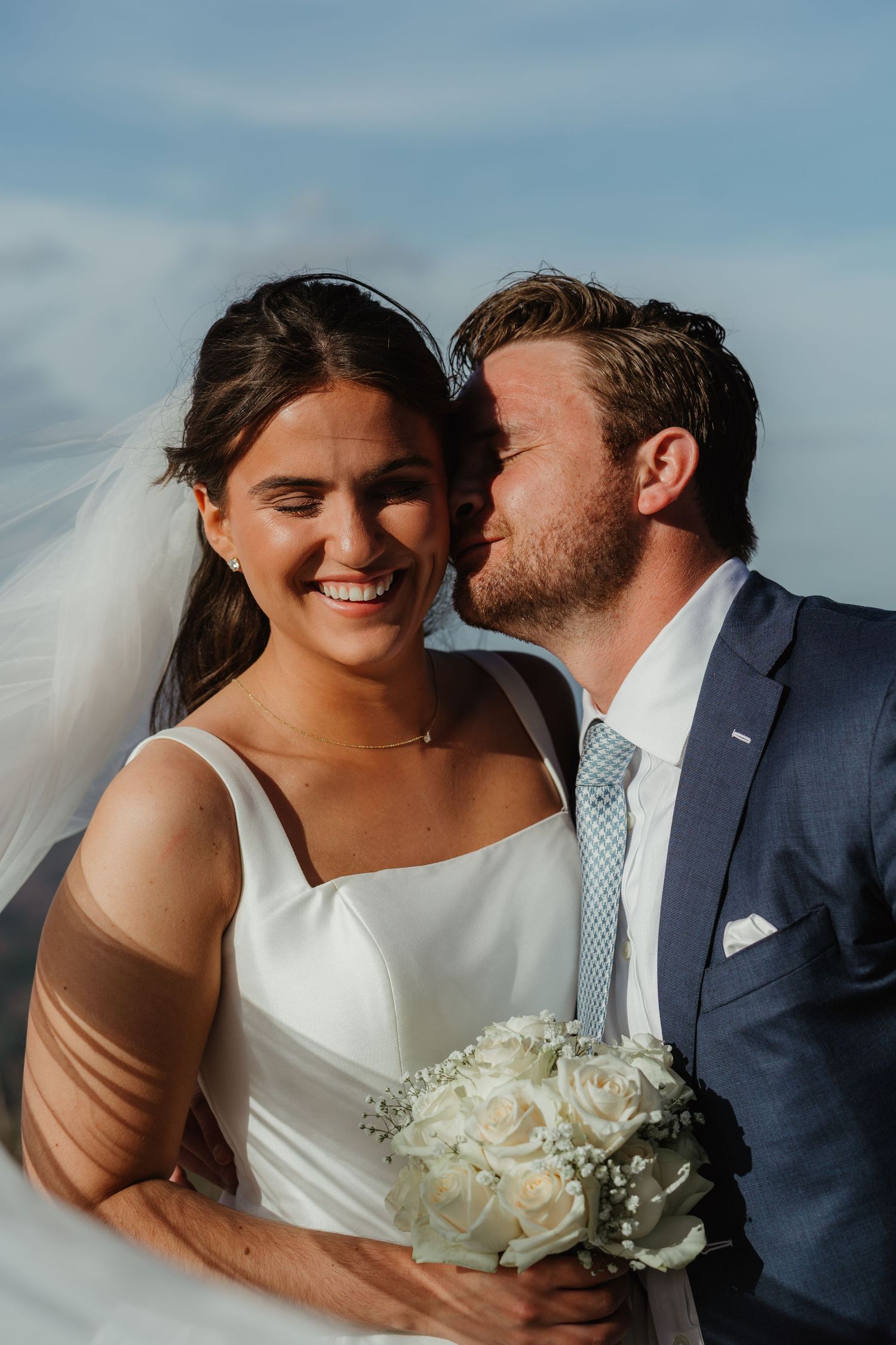 Groom in navy suit kisses smiling bride on the cheek as her veil blows in the wind under blue sky.