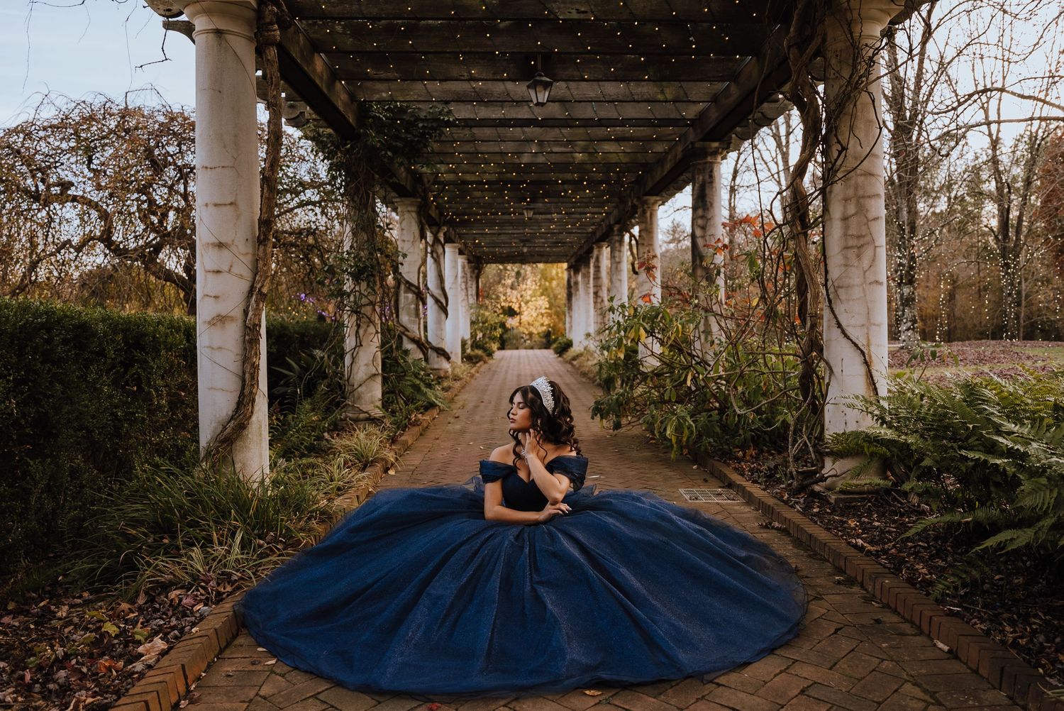 A young woman in a stunning navy blue ball gown poses along a pergola-lined garden pathway.