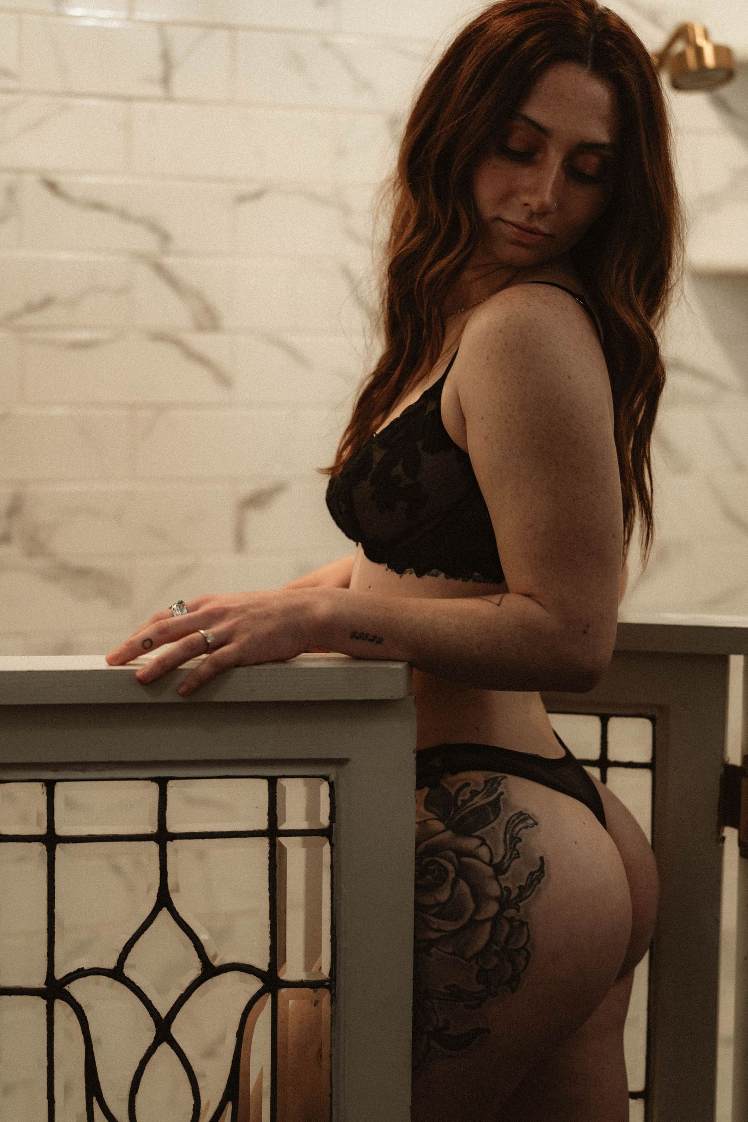 Woman in black lace lingerie with floral hip tattoo posing by vintage glass panel in marble bathroom.