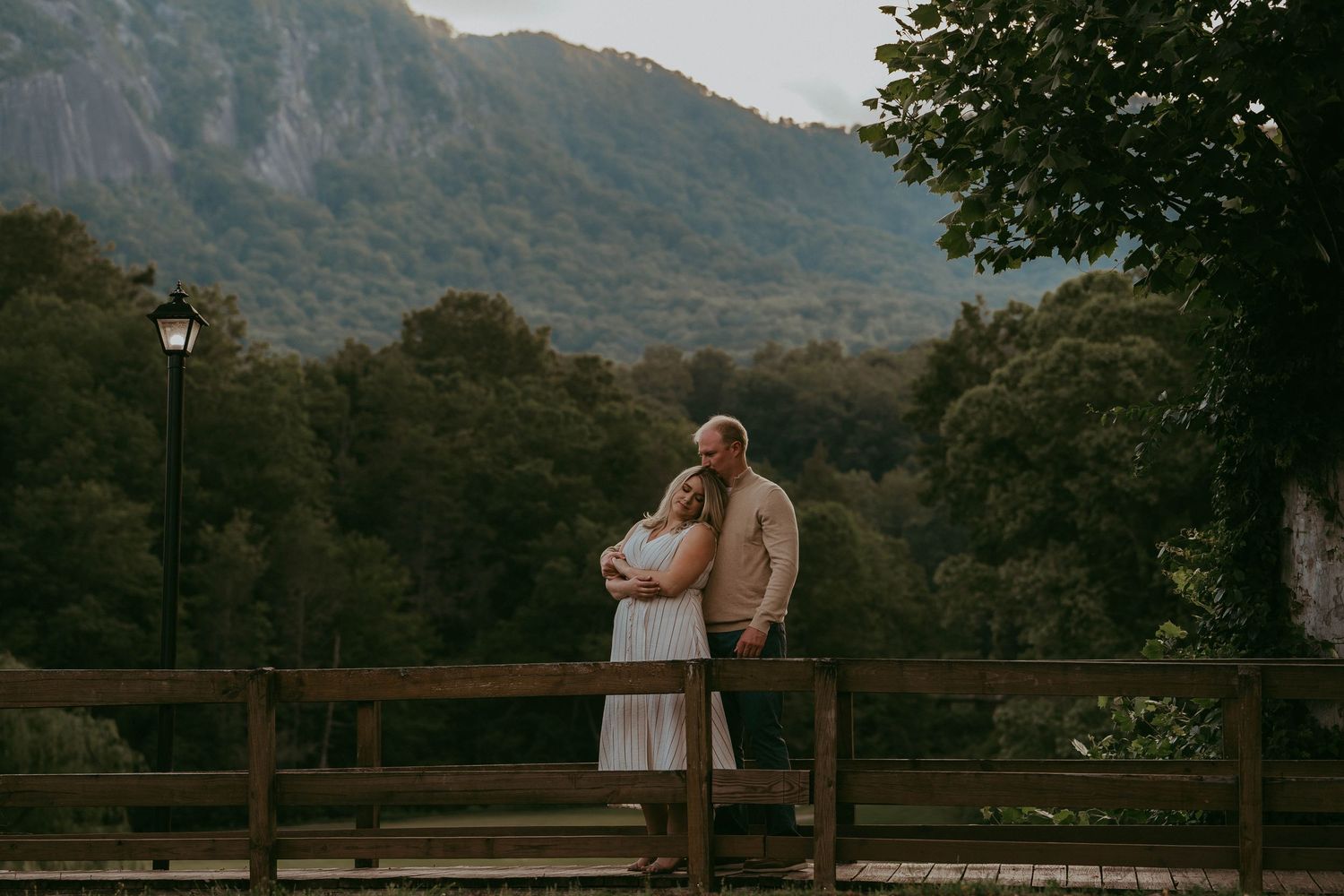 Romantic couple leaning in for a kiss by a wooden fence with mountain scenery behind.