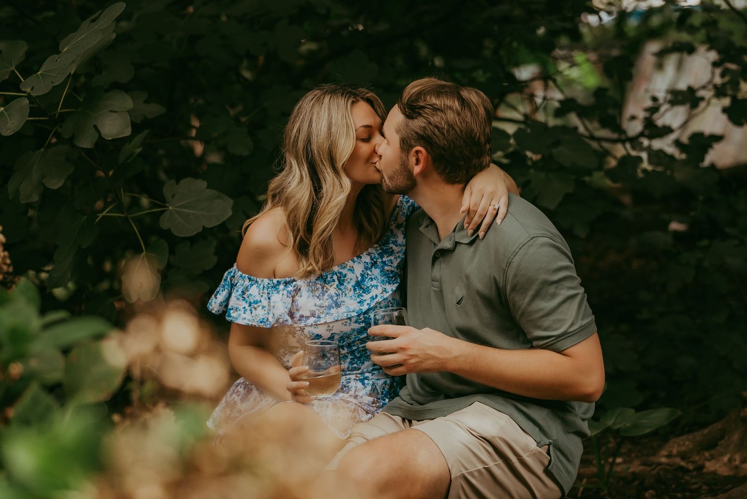 Couple kissing among lush green foliage, woman in blue floral dress holding a drink.