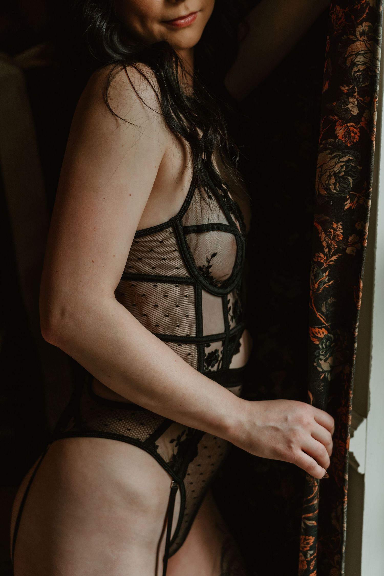 Woman in sheer black lace bodysuit and thong standing in a doorway with moody dramatic lighting.
