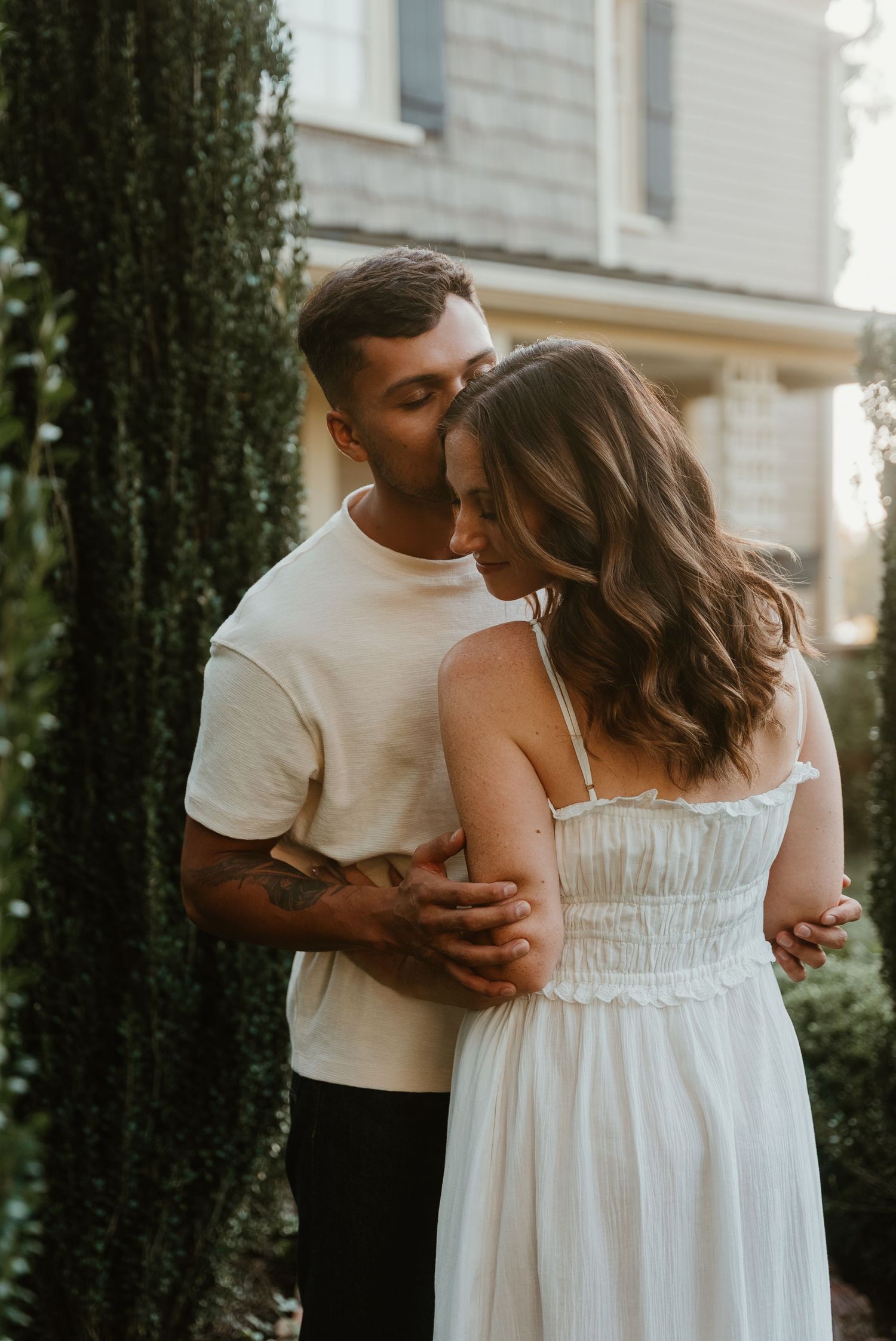 Man gently kissing woman on head while embracing her from behind near lush garden greenery.