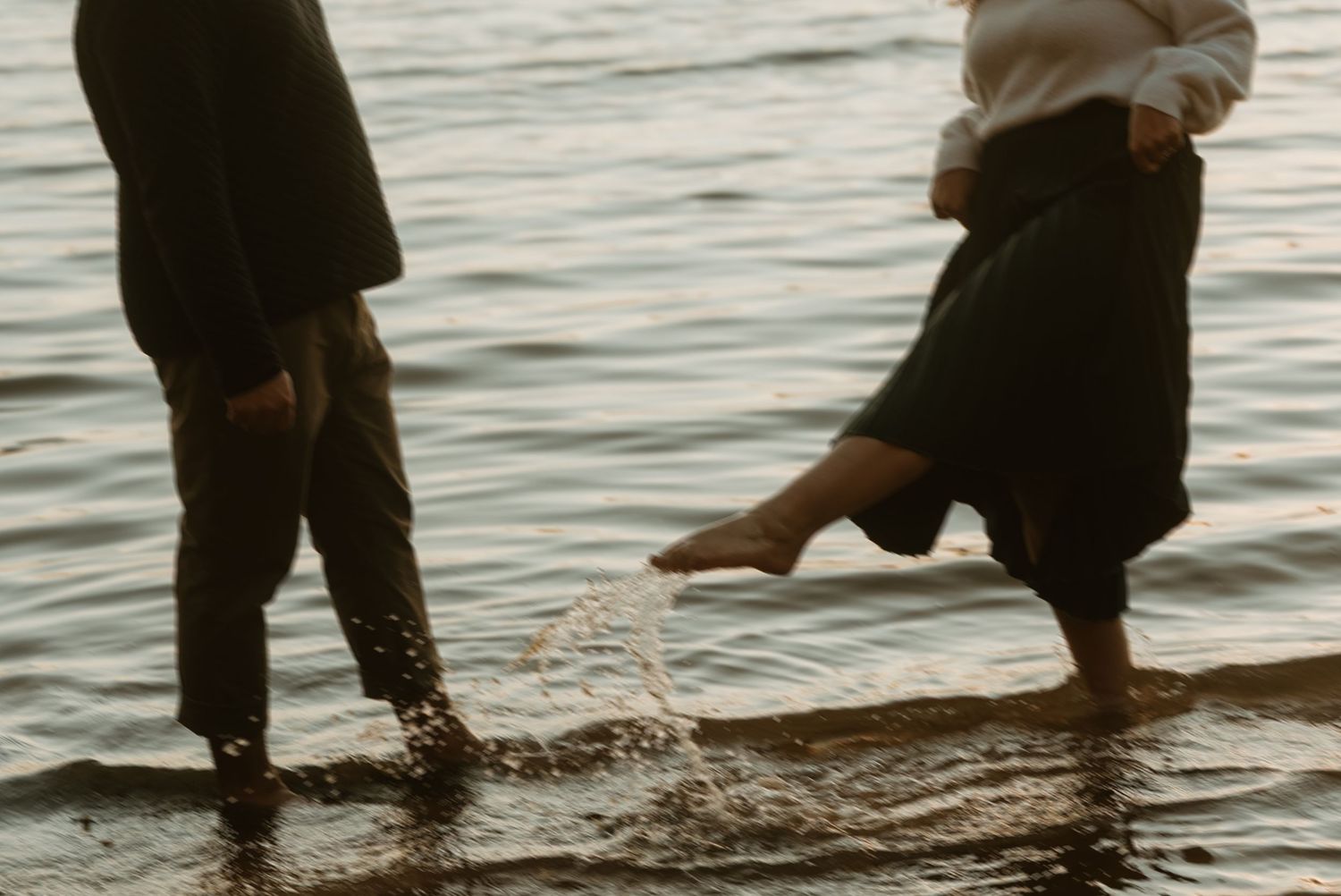 Couple splashing barefoot in shallow lake water at dusk, woman lifting her skirt.