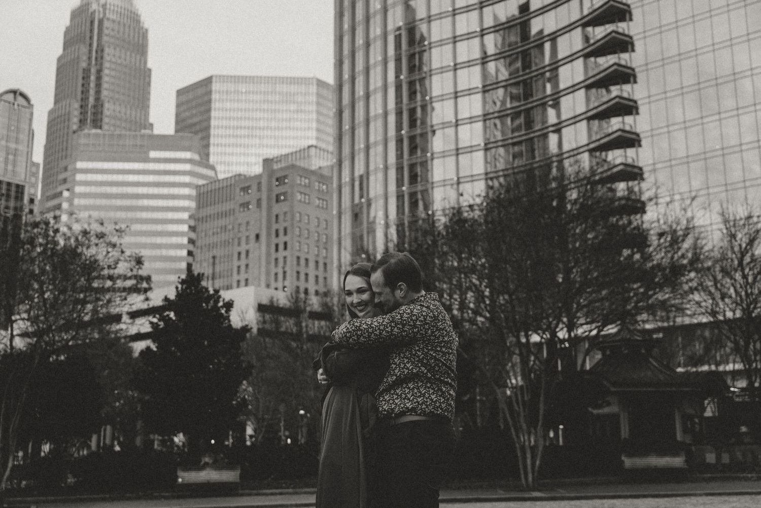 Black and white photo of a couple embracing in an urban setting with city skyscrapers in the background.