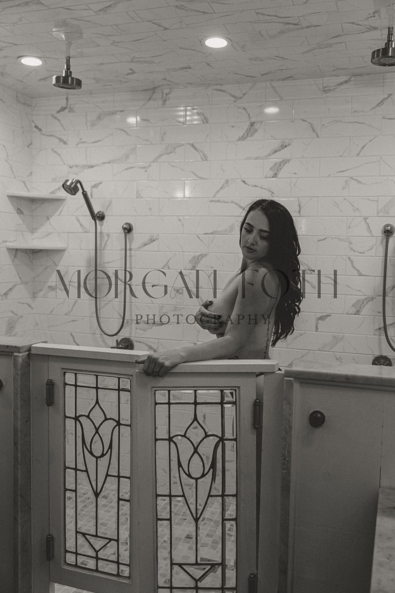 Black and white photo of brunette woman in marble shower with stained glass doors.