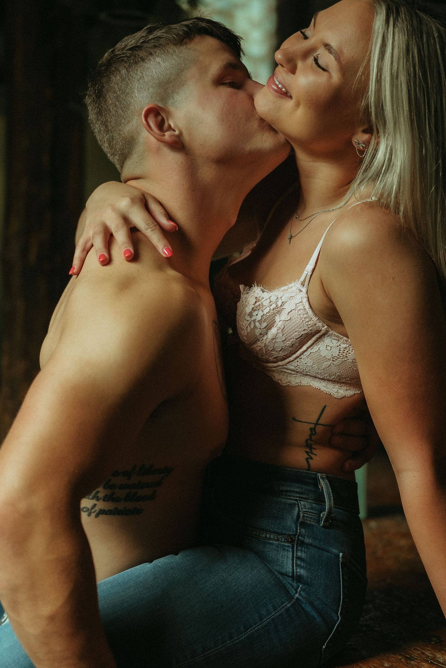 Tattooed shirtless man kissing smiling blonde woman in lace bralette in romantic couples boudoir session.