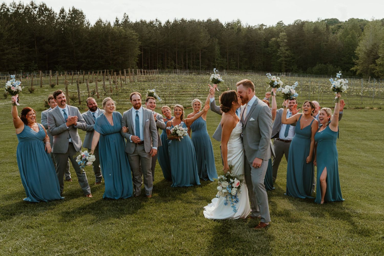 Wedding party celebrating with bride and groom kissing, bridesmaids in teal and groomsmen in gray suits.
