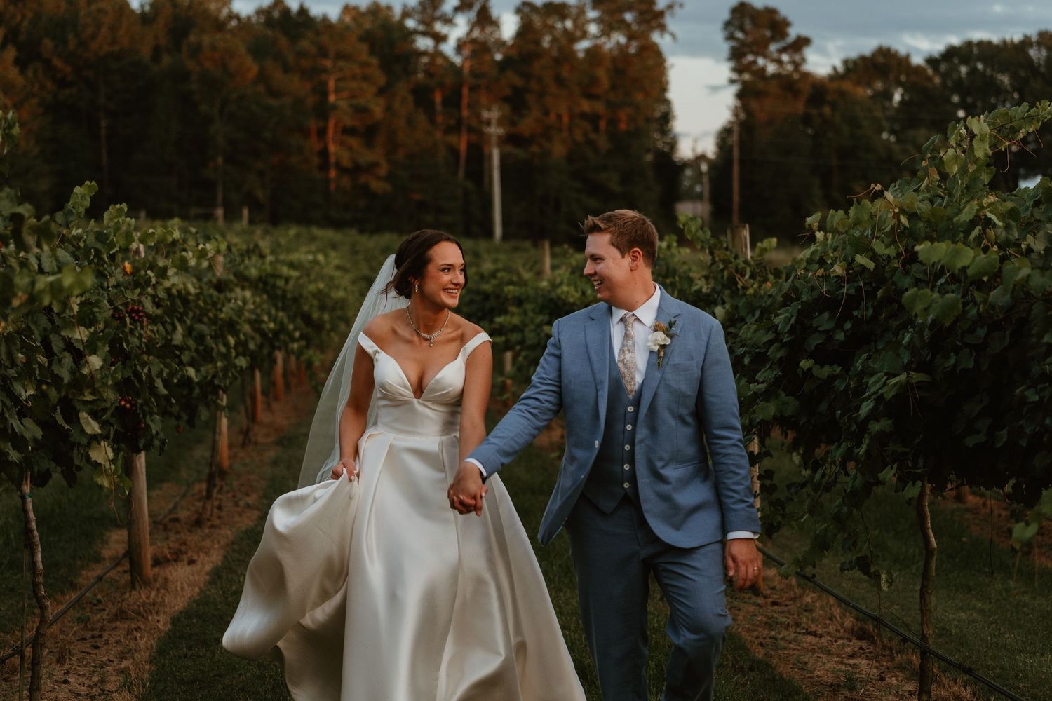 Bride and groom walking hand-in-hand through scenic vineyard rows in wedding attire at golden hour.