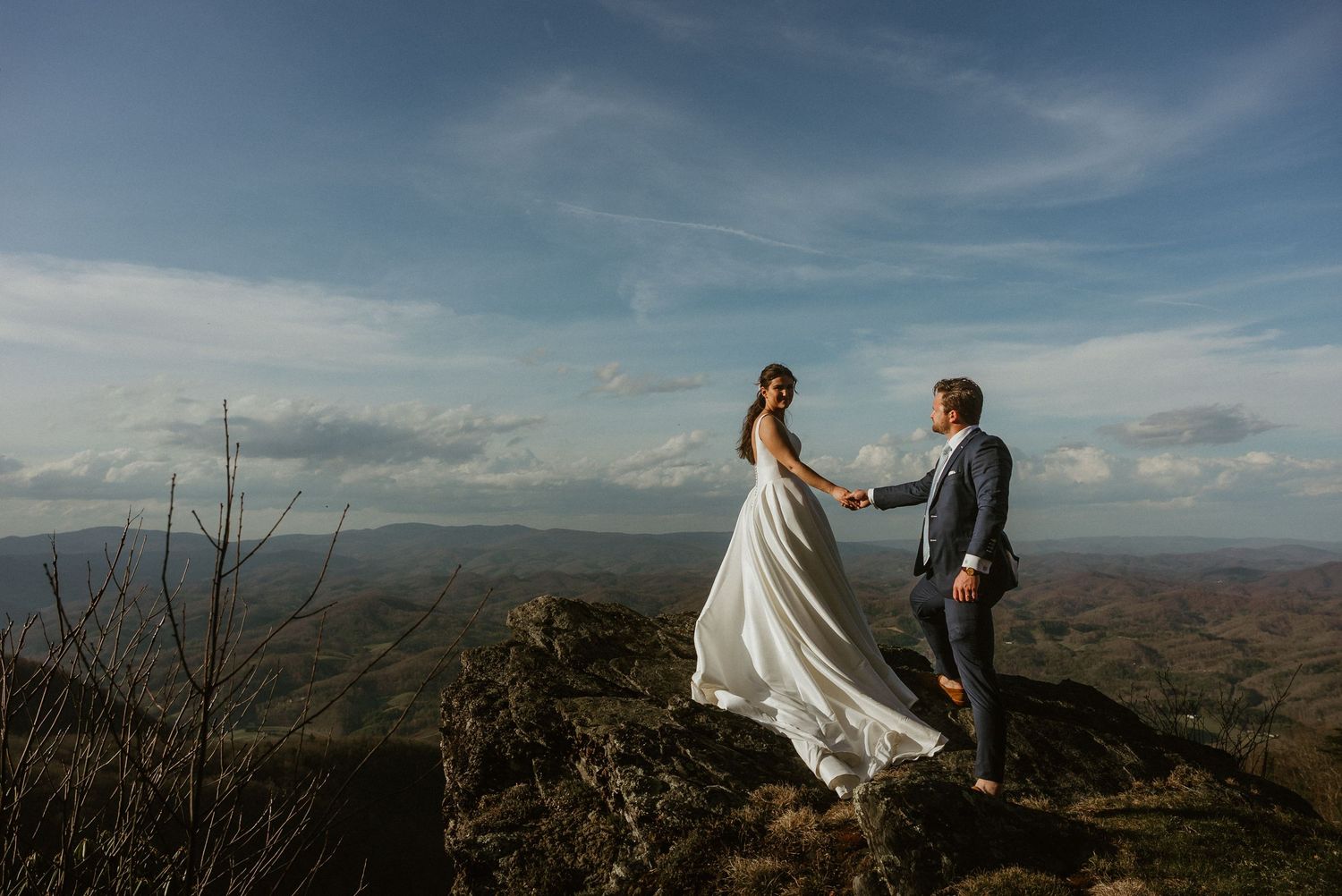 Bride and groom hold hands on a rocky mountain summit with sweeping valley views under a blue sky.