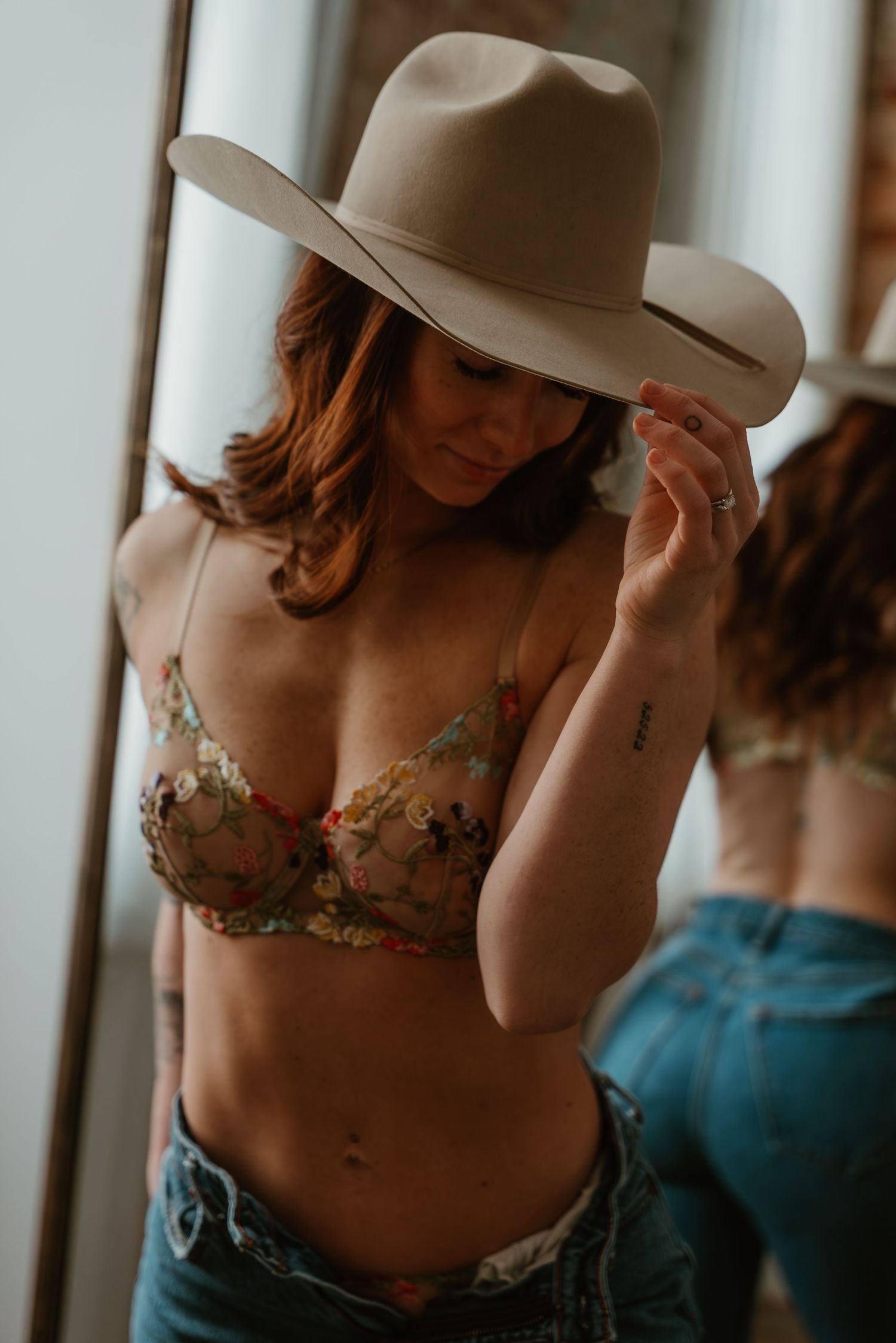 Woman in floral bra and jeans adjusting tan wide-brim hat while standing before mirror in soft light.