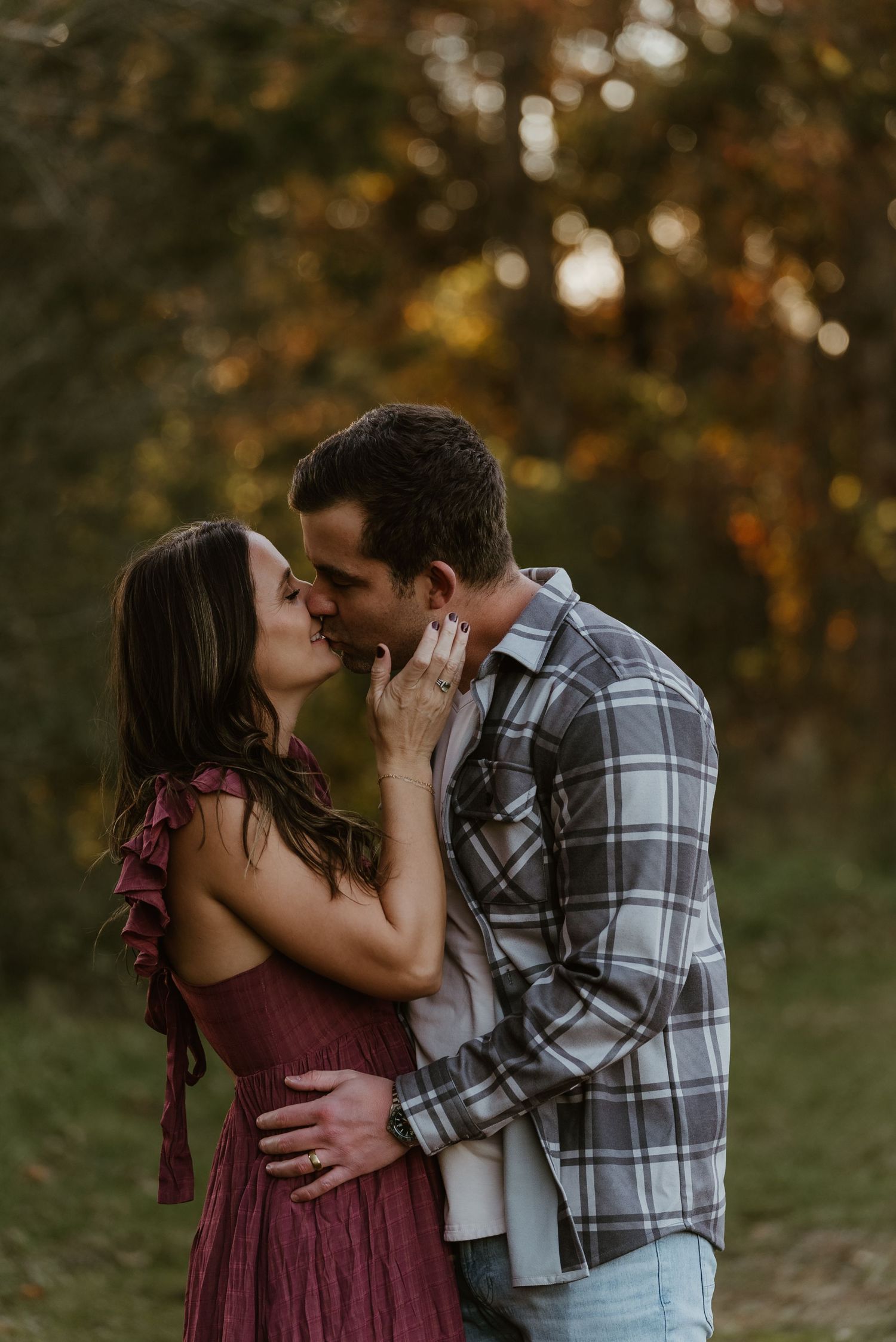 Romantic couple sharing a kiss outdoors in a forest setting at golden hour, woman in red dress.